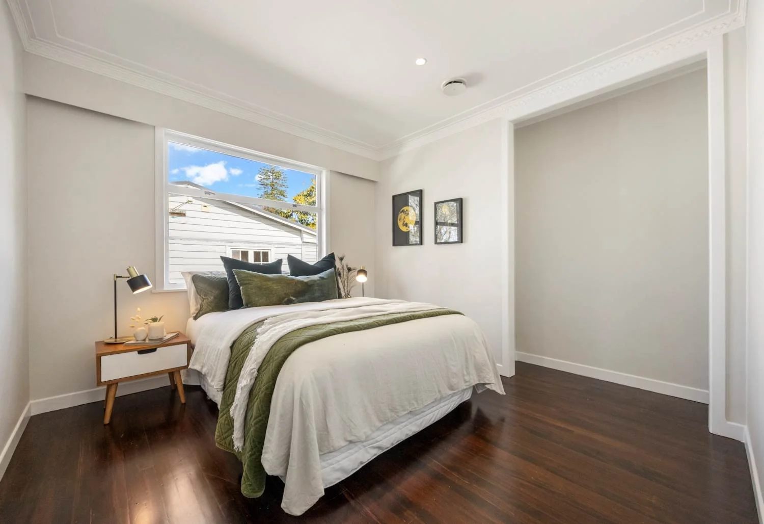 Bright bedroom with white walls, dark hardwood floor, bed with white, green, and gray bedding, wooden nightstand with black and gold lamp, three framed pictures on wall, and window showing exterior of neighboring house and blue sky.