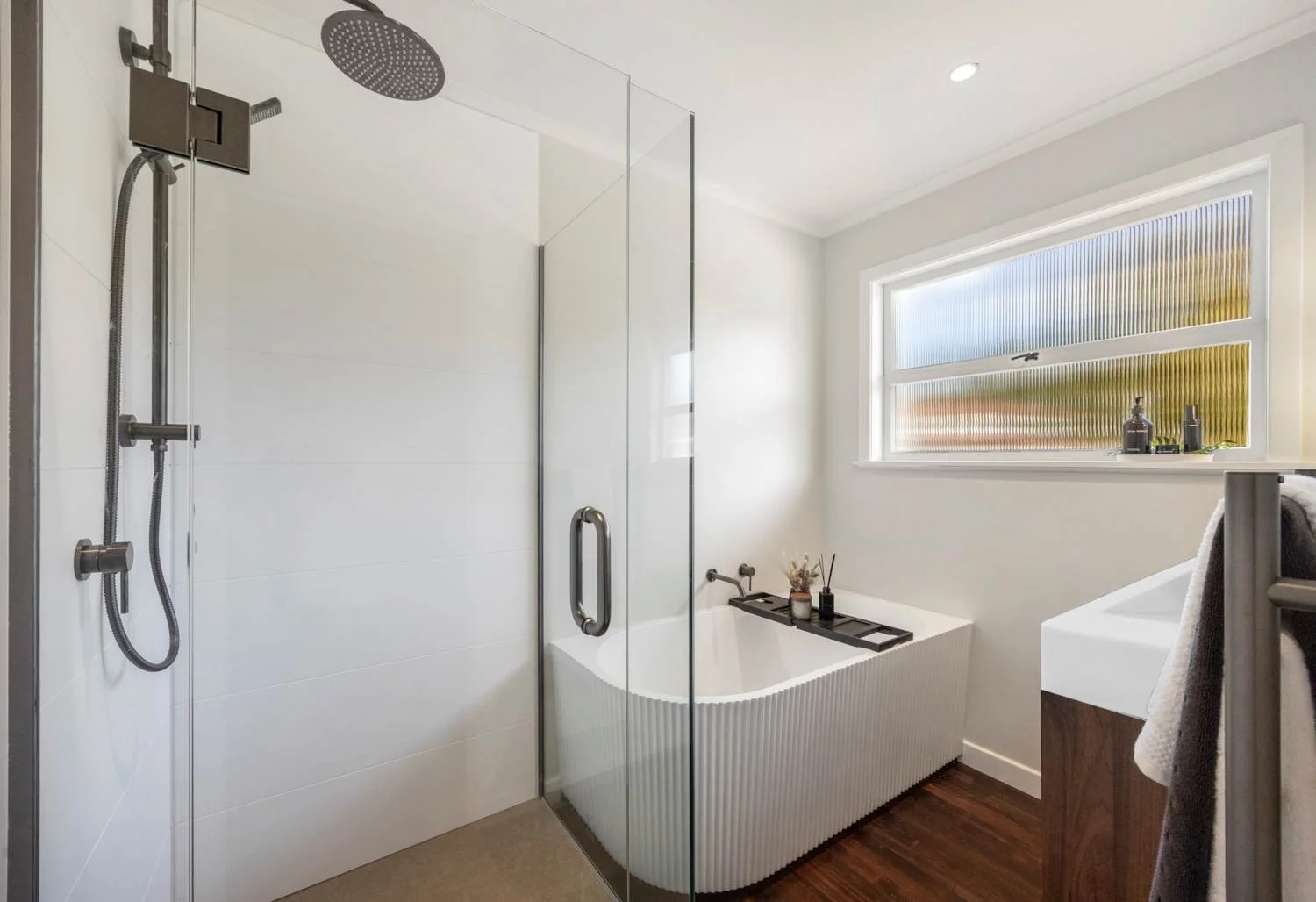 Modern bathroom featuring a walk-in shower with a rainfall showerhead, a freestanding bathtub with a black tray, a window with a frosted glass pattern, and a wooden vanity with a towel hanging on a rack.