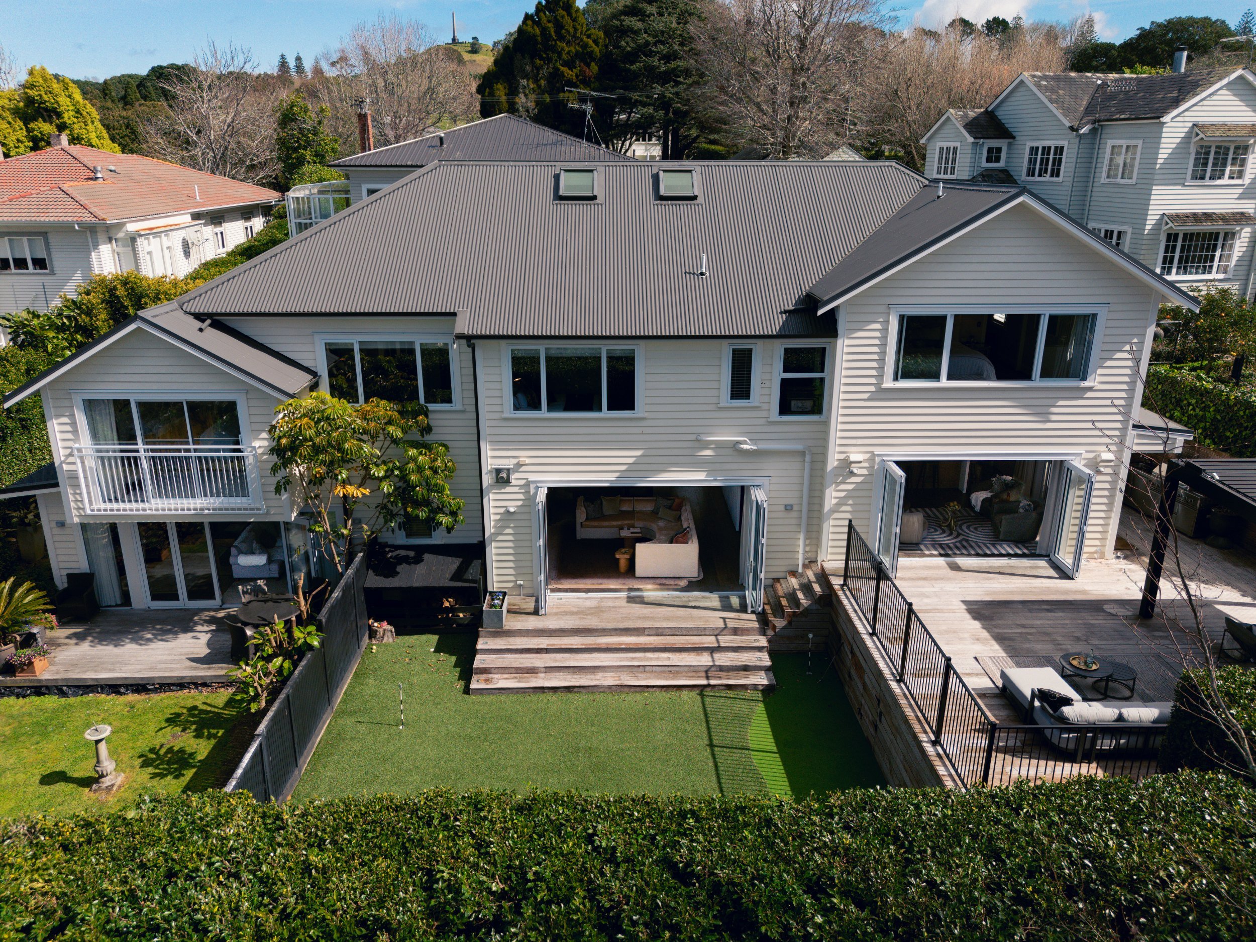 An aerial view of a residential backyard showing a deck with outdoor furniture, green lawn, trees, and neighboring houses with tiled and metal roofs.