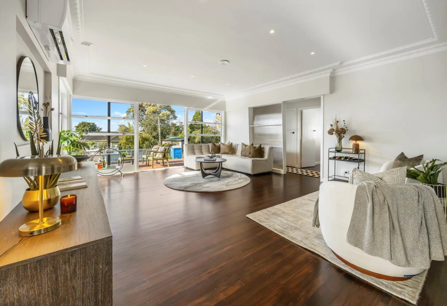 Bright living room with large windows, white walls, dark hardwood floors, beige and white furniture, a round rug, plants, and decorative accents.