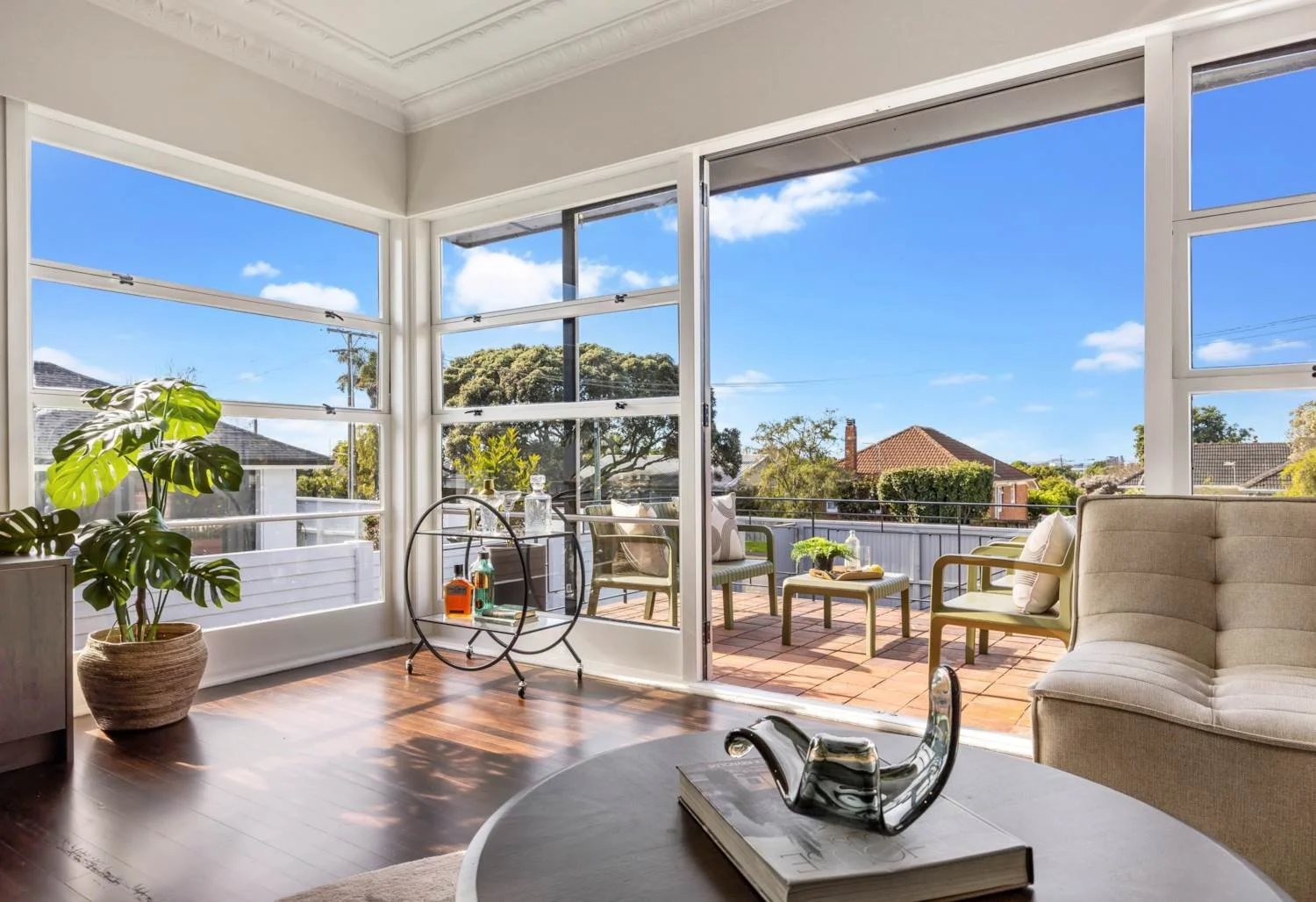 Sunlit living room with large windows opening to a balcony with outdoor chairs and greenery, a potted plant on the floor, a beige couch, and a coffee table with a decorative tray and books.