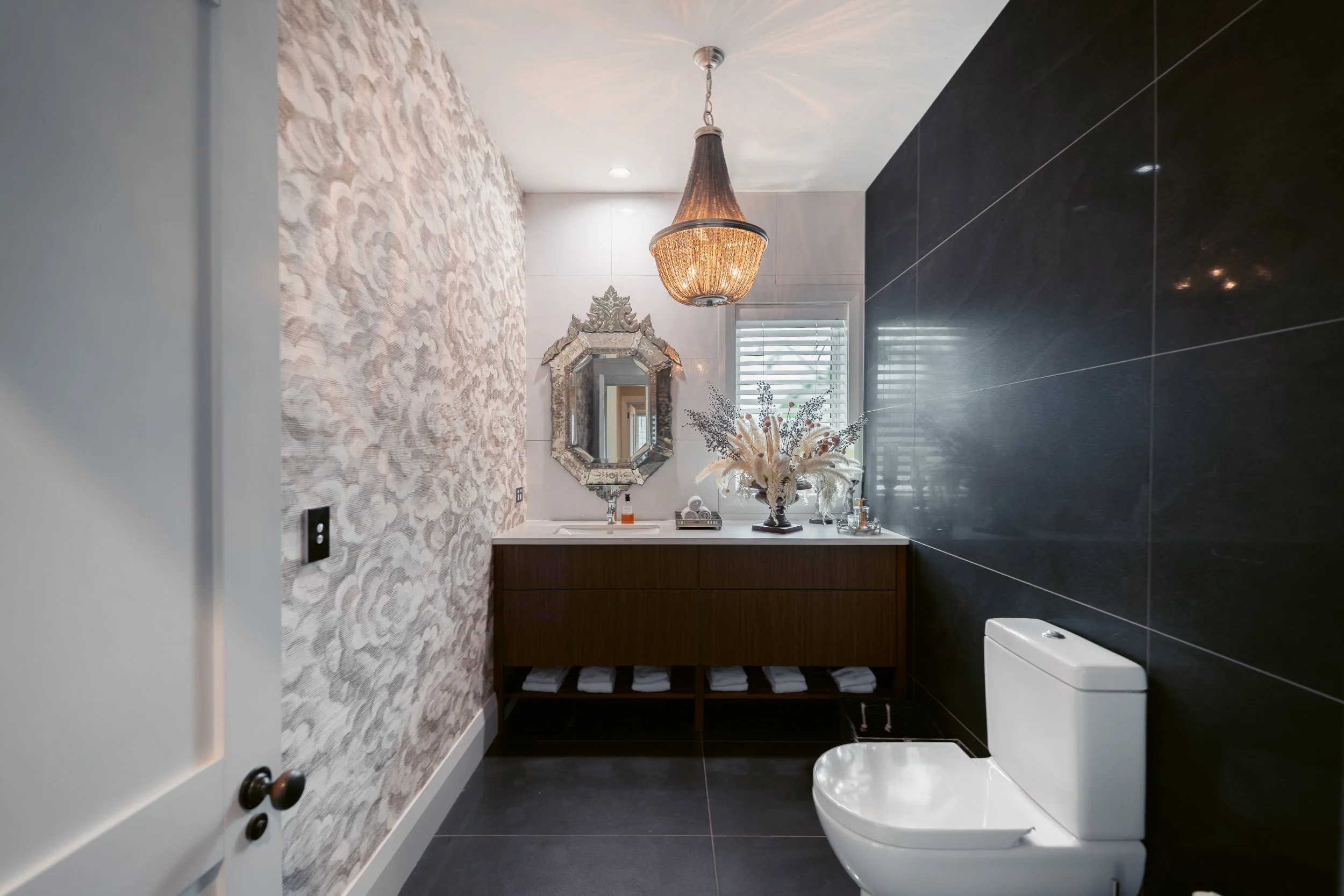 Modern bathroom with black tiled walls, a white toilet, a wooden vanity with folded towels, a decorative mirror, and a large floral arrangement, illuminated by a statement chandelier and natural light from a window with blinds.