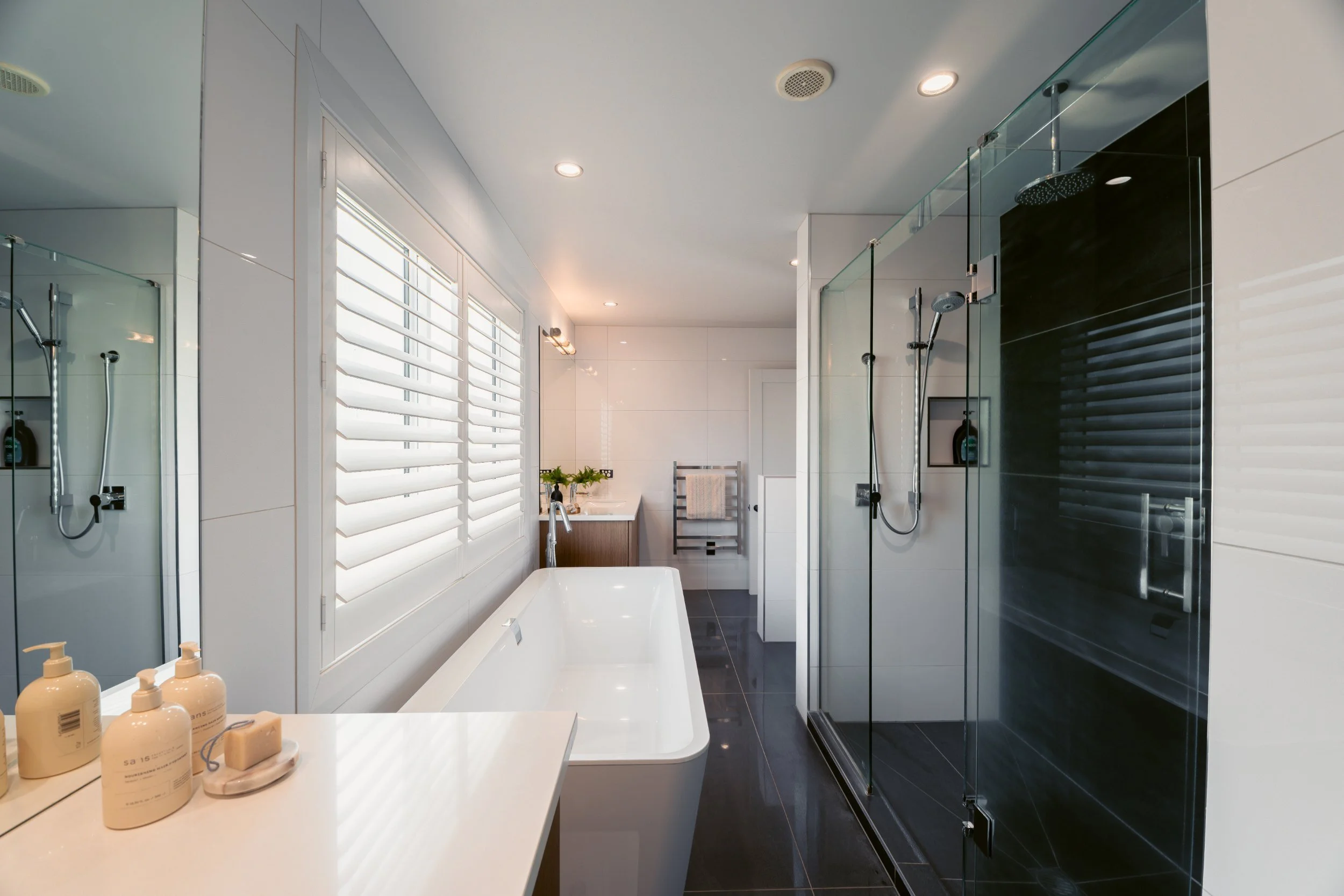 Modern bathroom with a white bathtub, black tiled shower, and large windows with plantation shutters.