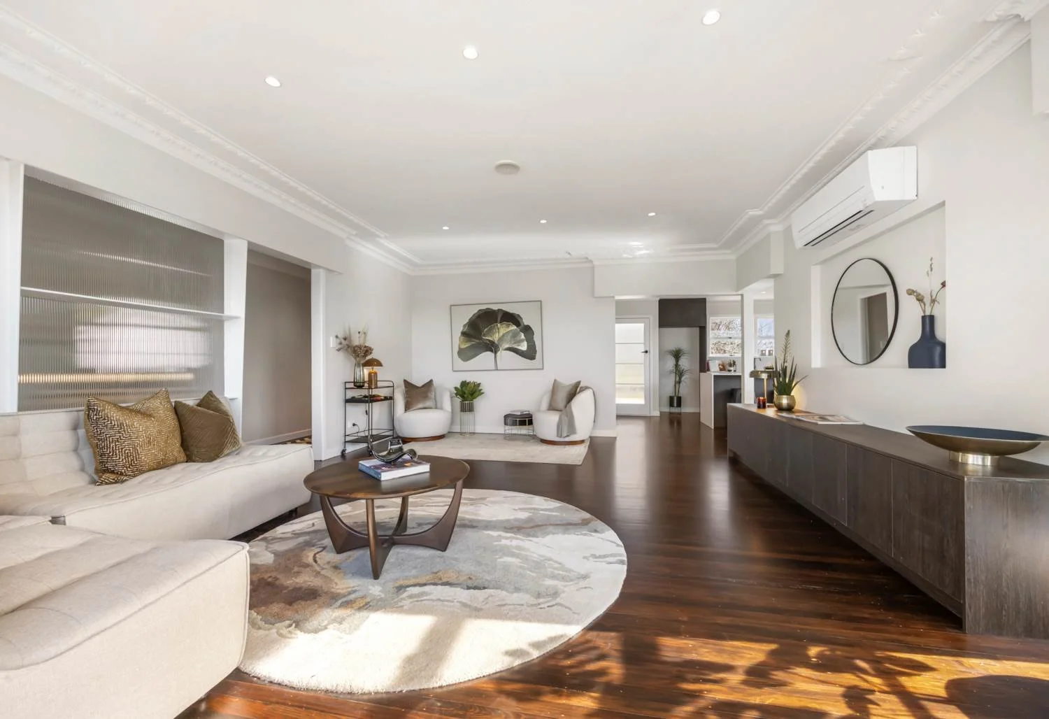 Bright and modern living room with white walls, dark wood floors, and contemporary furniture. Features a large beige sofa with decorative pillows, a round wooden coffee table, and an area rug. Two white armchairs with pillows are placed against the w