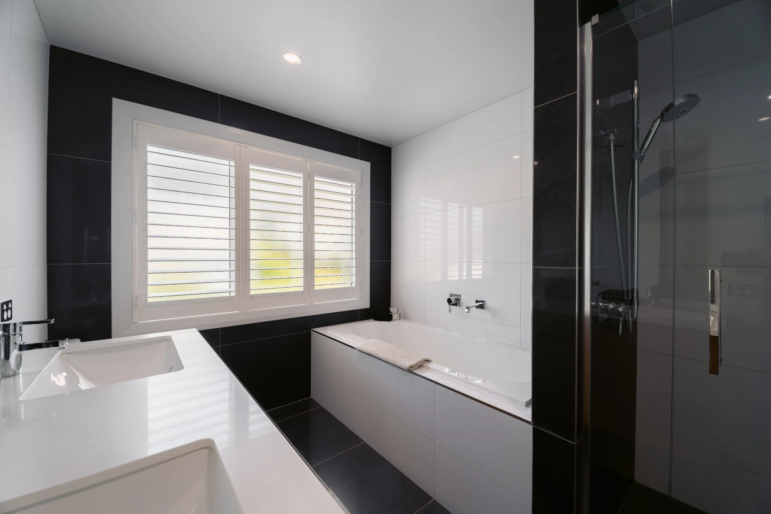 A modern bathroom with a white bathtub, black and white tiled walls, a large window with white shutters, and a glass shower enclosure.