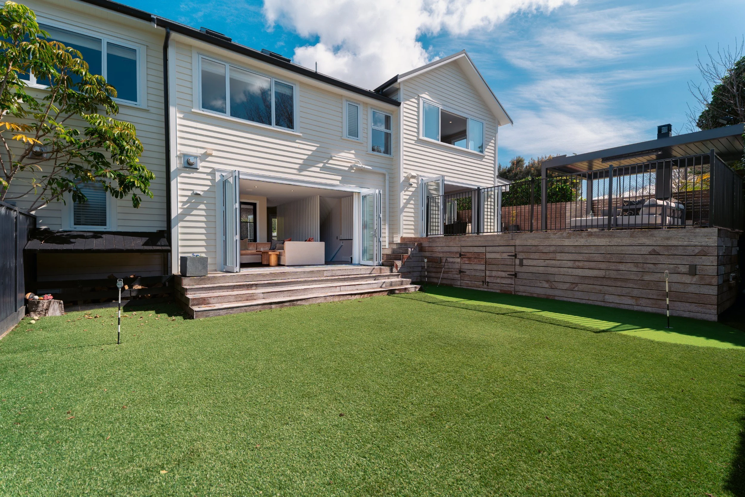 Backyard with green artificial grass, wooden stairs leading to a patio, and a white two-story house with large windows, partly open patio doors, and a fenced outdoor seating area beneath a covered deck.