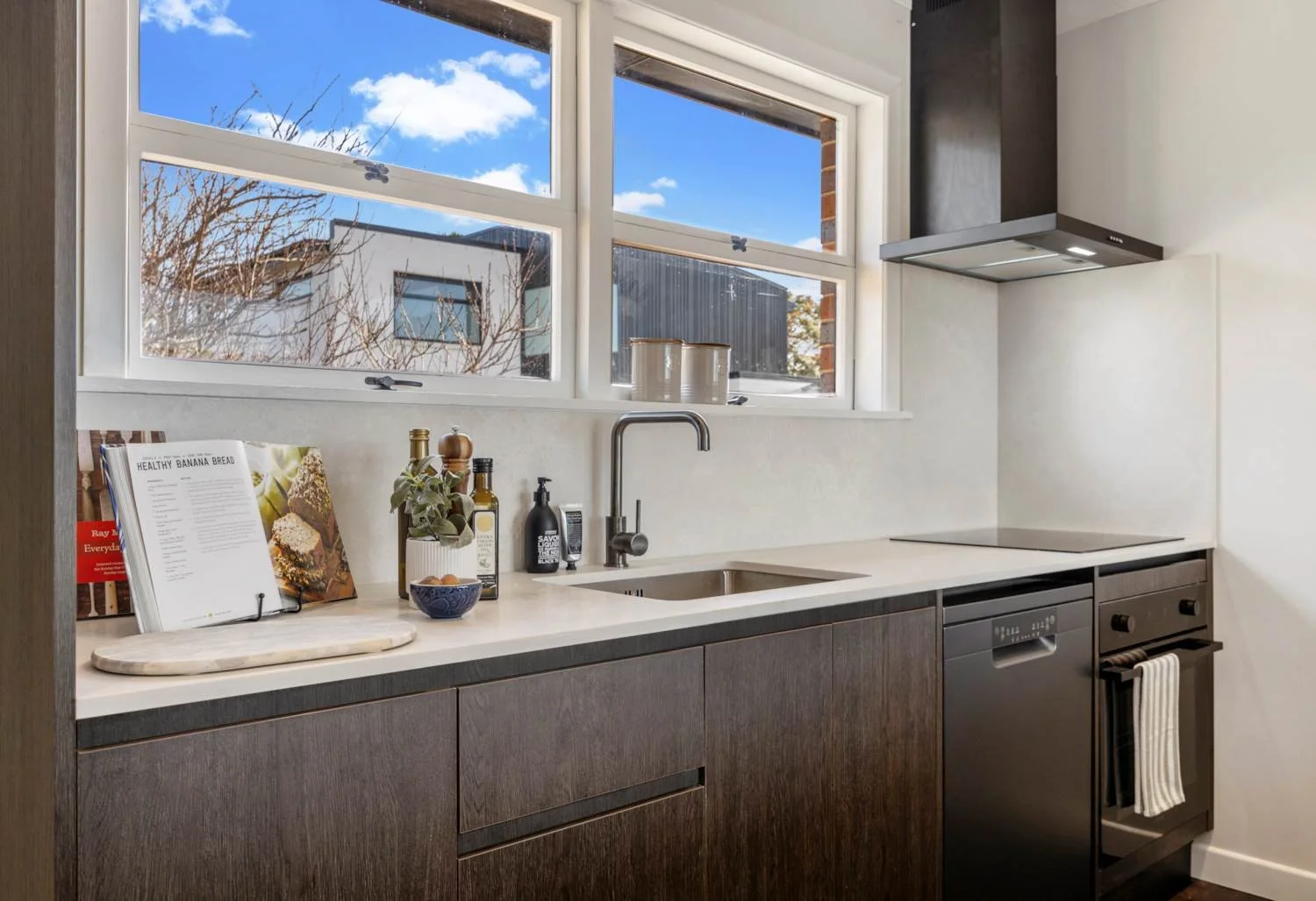 Modern kitchen with dark wood cabinets, white countertop, and large windows showing a blue sky with some clouds. There are items such as cookbooks, bowls, bottles, and a plant on the counter, a stove with a towel hanging, and an integrated dishwasher
