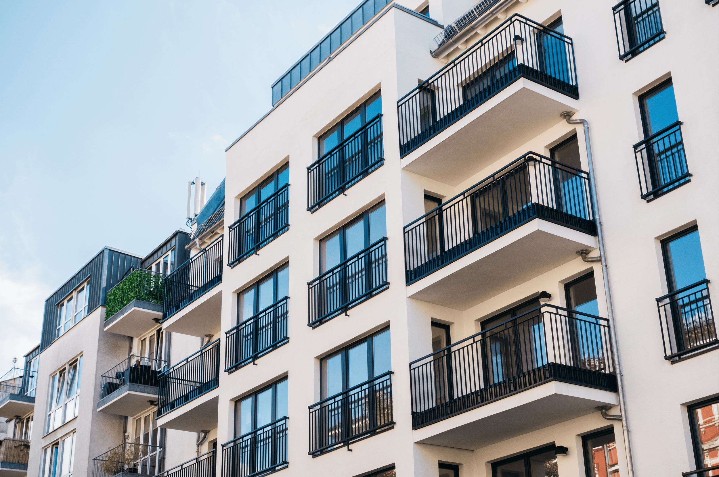 Modern white apartment building with multiple floors, each featuring black metal rail balconies and large windows, with a clear blue sky in the background.