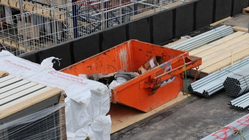 Self Dumping Trash bin in orange on a construction job site in the United Kingdom. The bin is partially filled with waste.