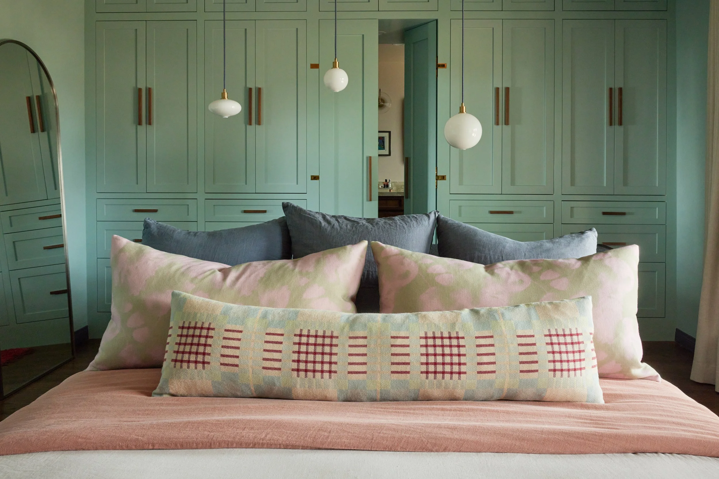 A bedroom with a bed in the foreground covered with a pink bedspread and multiple pillows, including one with a pastel checkered pattern. Behind the bed, there is a large built-in mint green wardrobe with wooden handles and a small open doorway revealing a bathroom with a mirror and framed picture. Three pendant lights hang from the ceiling, illuminating the room.