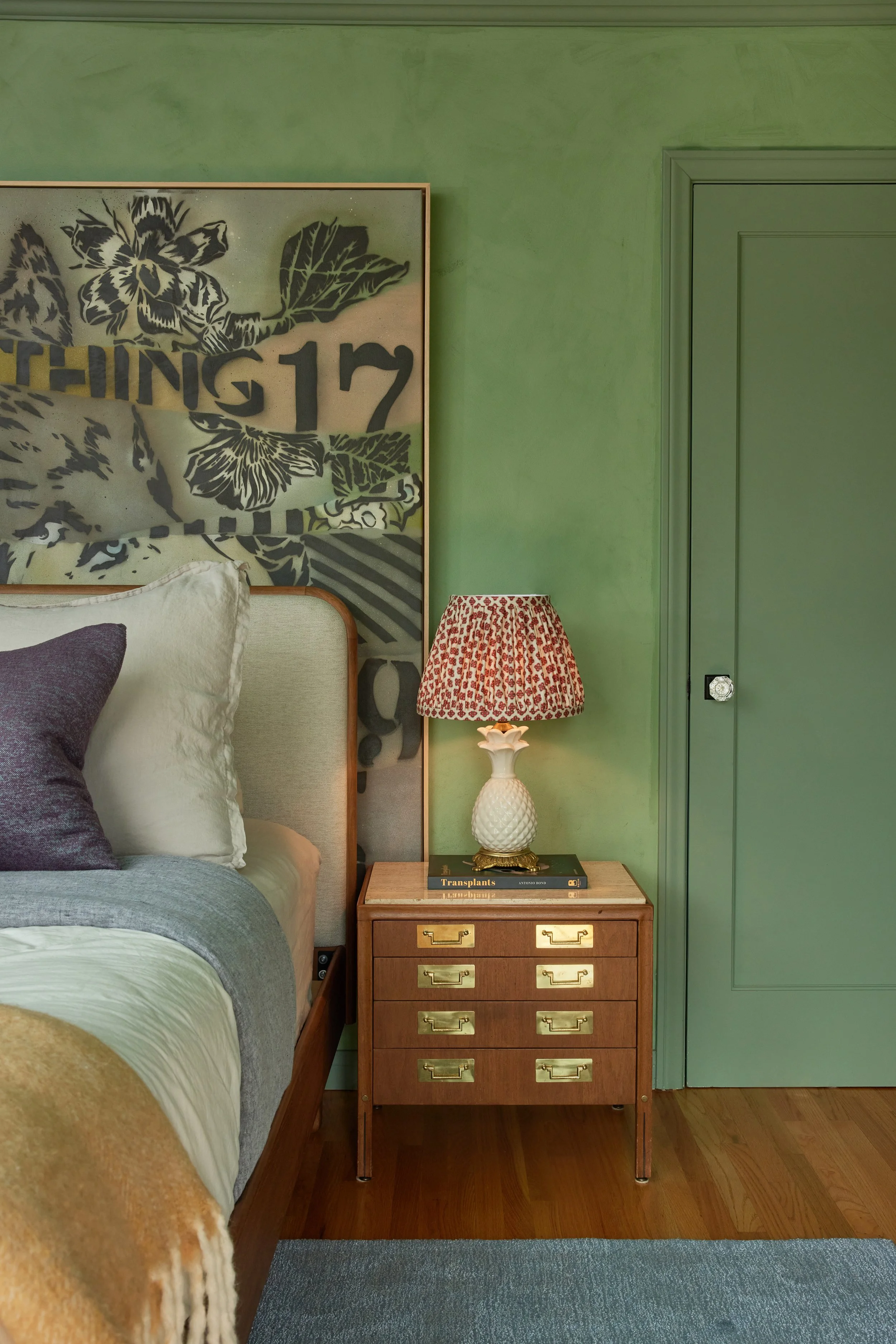 A bedroom corner featuring a bed with pillows, a vintage wooden nightstand with brass handles, a pineapple-shaped lamp, a few books, a green wall with a framed artwork of black floral and text, and a blue and green rug on a wooden floor.