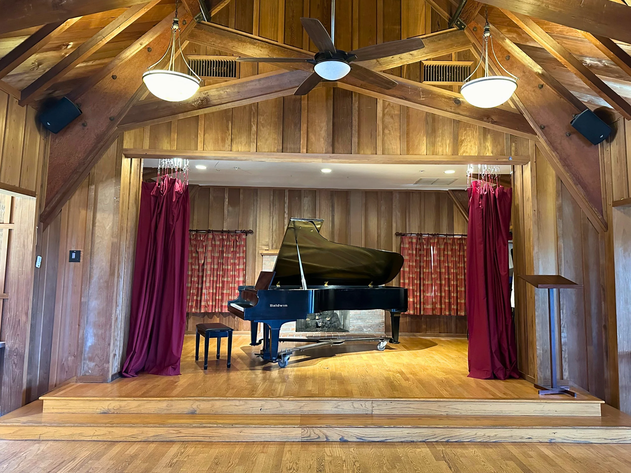 Wood-paneled stage with a black grand piano, red curtains, and a stool in a cozy performance room.
