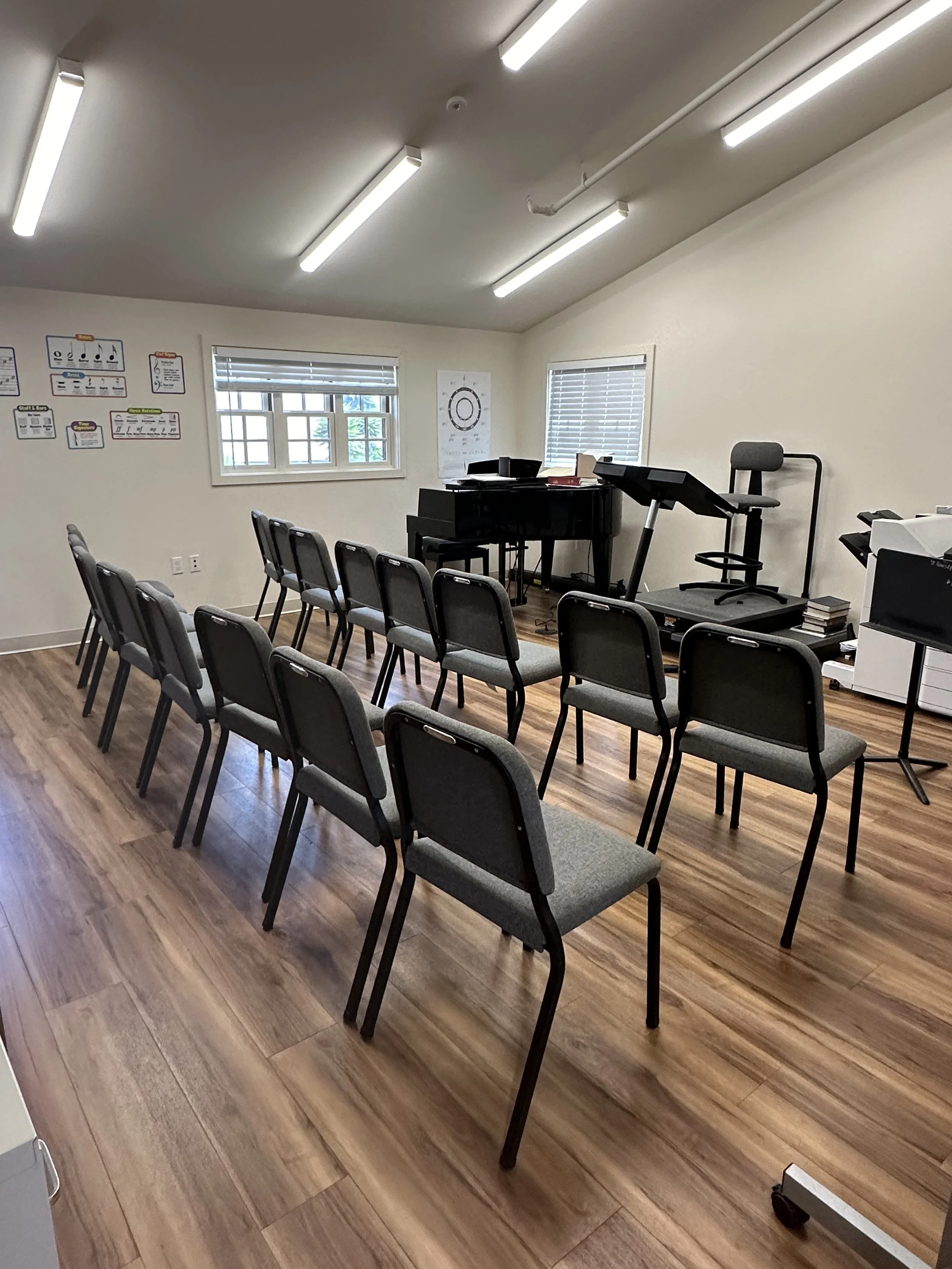 An empty classroom or meeting room with chairs arranged in rows, a piano, a standing desk, and posters on the wall.