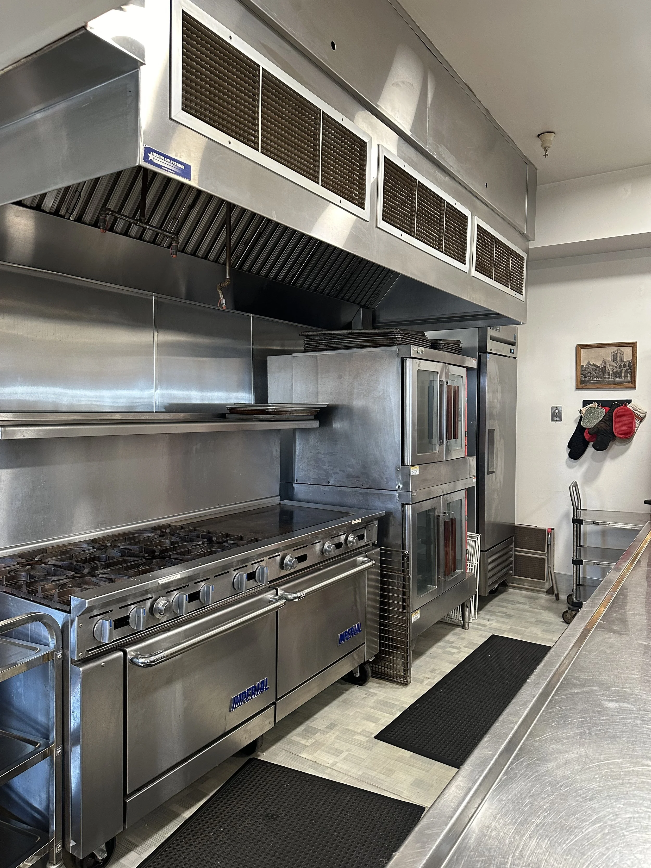 Commercial kitchen with stainless steel stove, oven, and ventilation hood, along with shelves, mats, and wall-mounted oven mitts.