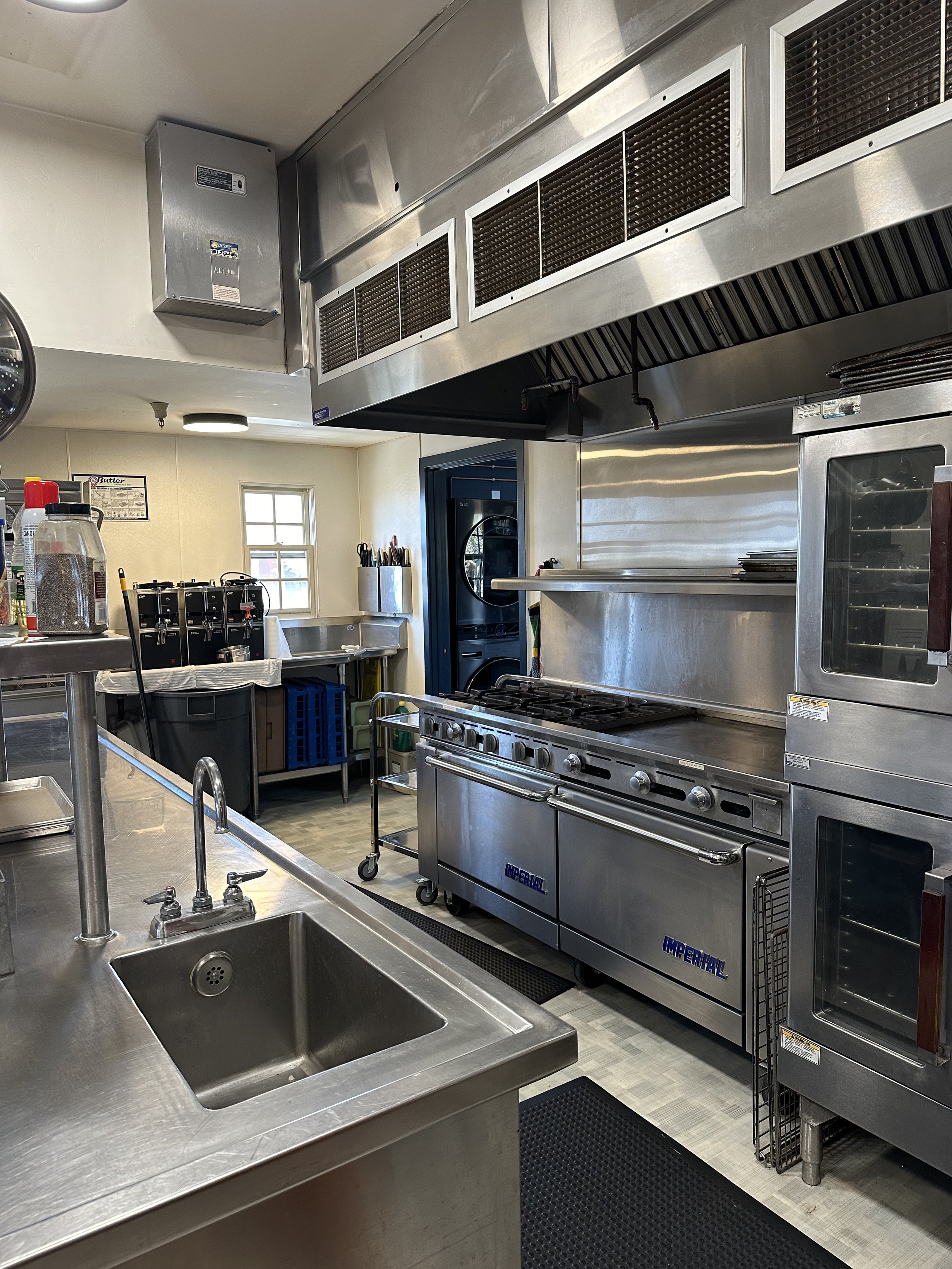 Commercial kitchen with stainless steel appliances, a sink, stove, oven, and various utensils. Window and cleaning supplies visible in the background.