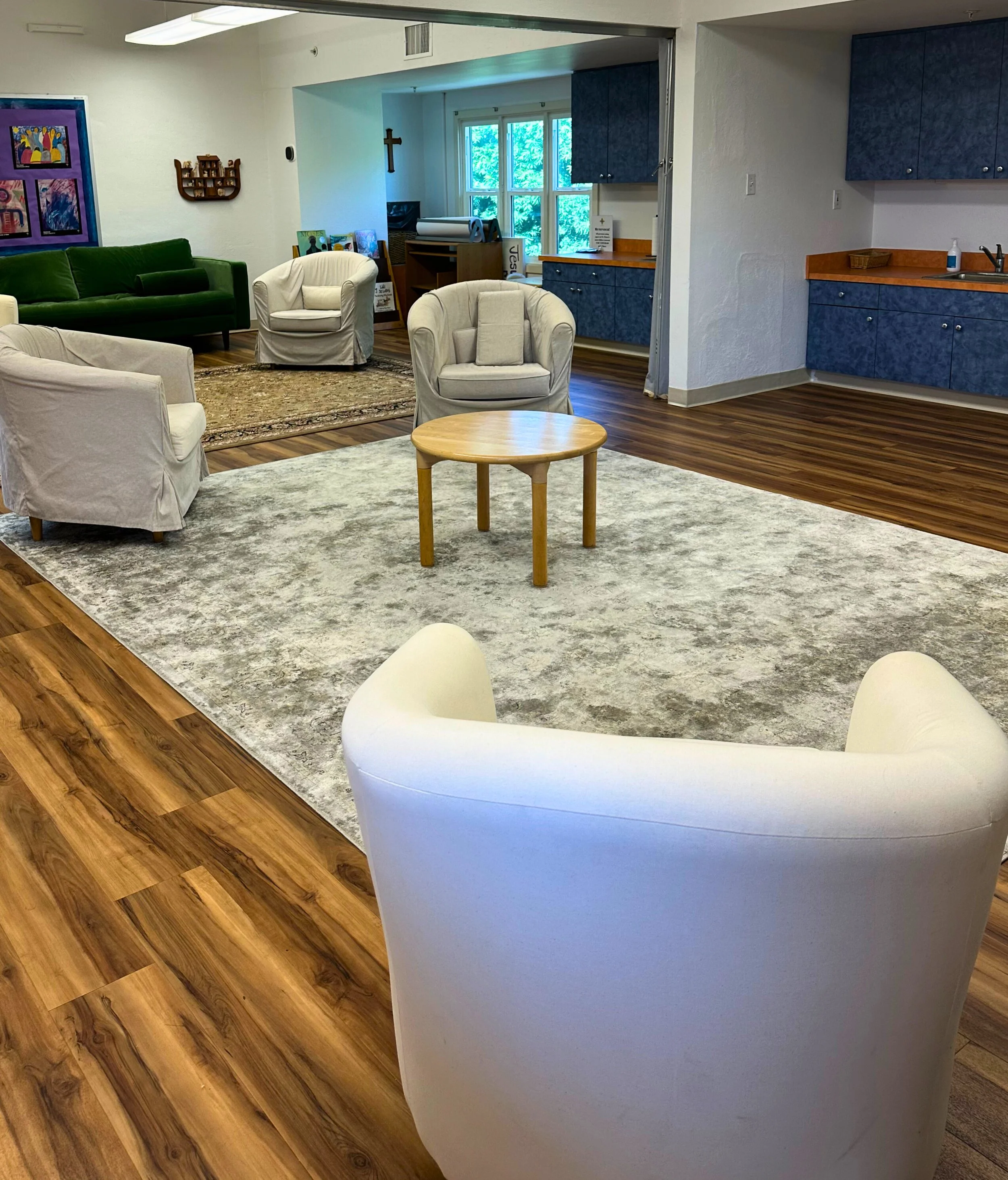 A cozy lounge area with multiple cream-colored armchairs, a green sofa, and a wooden coffee table on a large beige and gray rug, with blue cabinets and a window in the background.