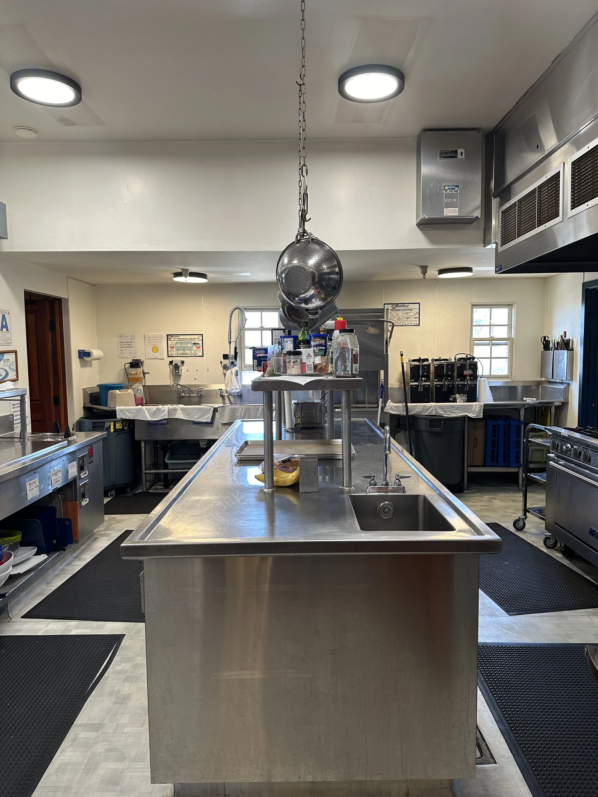 Empty commercial kitchen with stainless steel island, hanging metal ladle, kitchen utensils, and various appliances and tools in background.