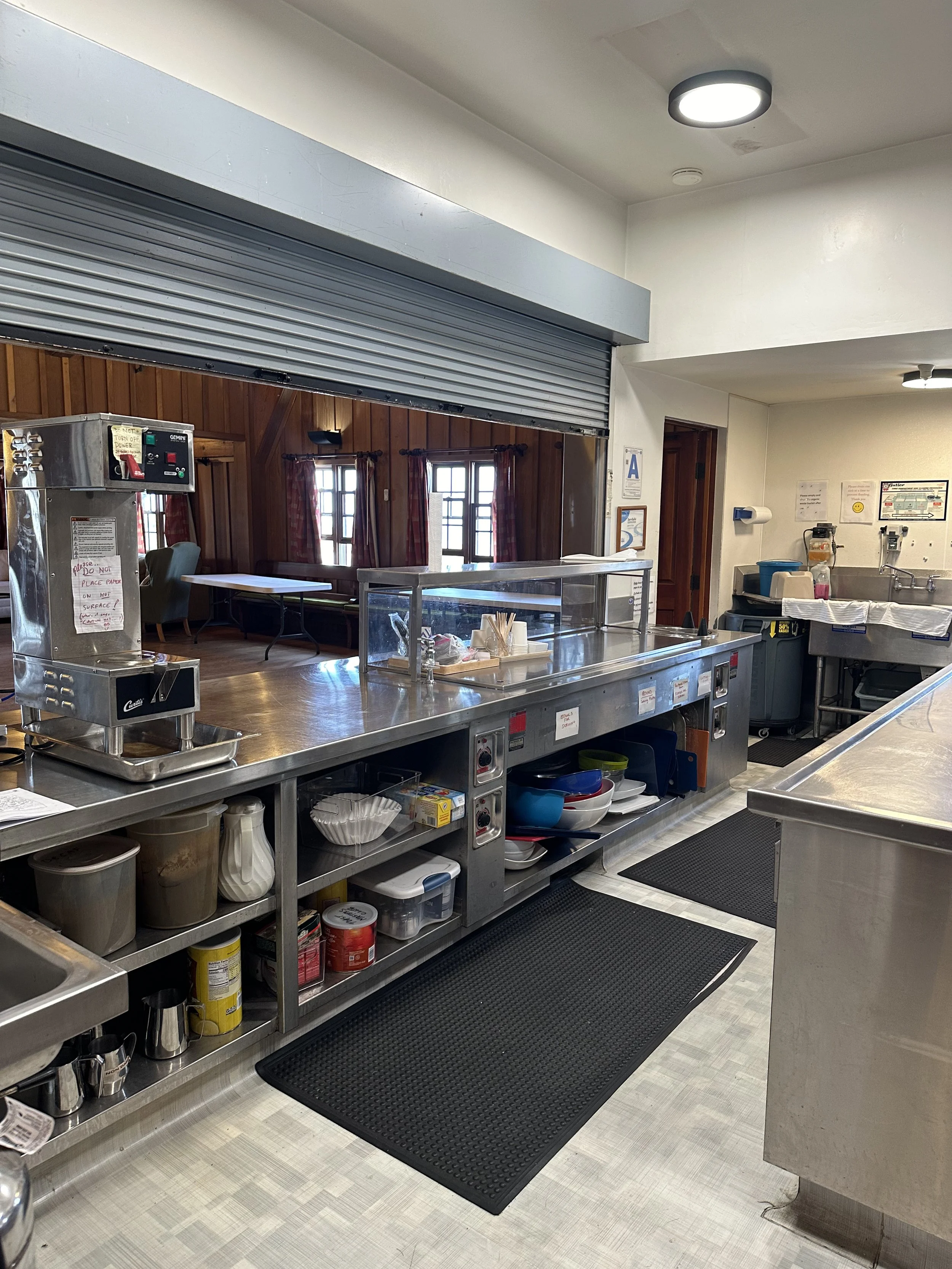 Empty commercial kitchen with stainless steel counters, sinks, and shelving, black rubber mats on the floor, and a partially open metal roll-up door leading to a dining area with wooden walls and windows.