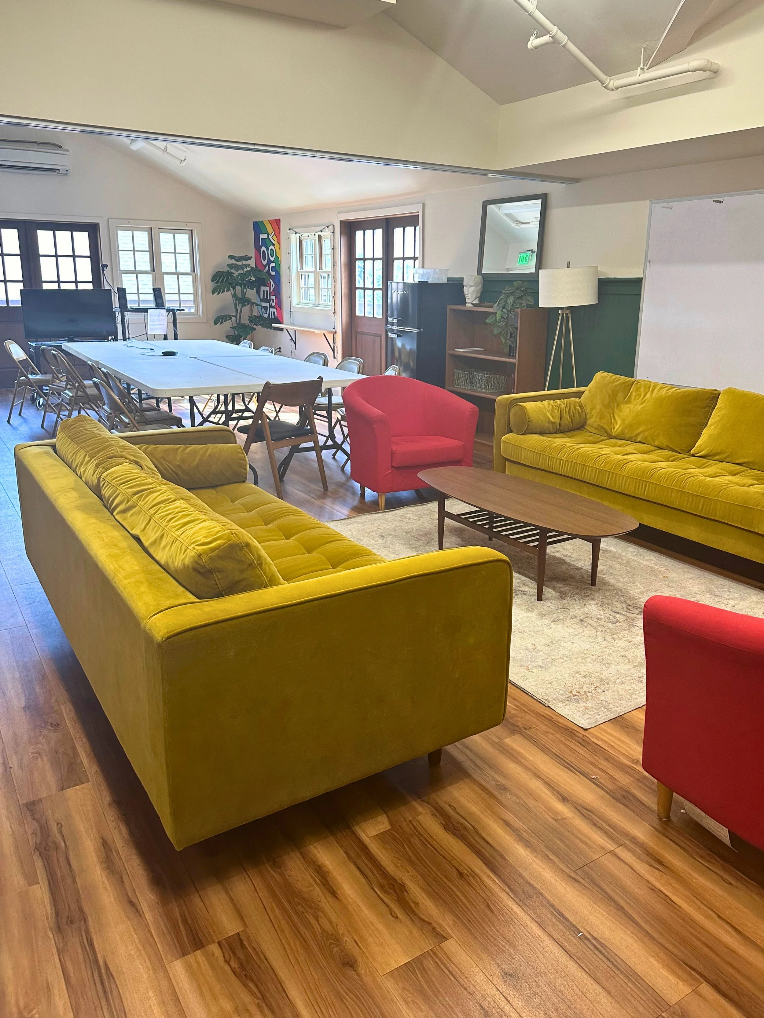 Living room with yellow and red sofas, a wooden coffee table, a large whiteboard, and a dining area with a long table and chairs, near windows and a door, with a rainbow-colored banner and a plant.