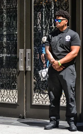 A security guard standing outside a building with black iron doors and decorative glass. He is wearing a black polo shirt, dark pants, sunglasses, and has a watch on his wrist.