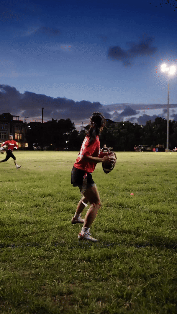 A person in a red shirt playing flag football on a grassy field at dusk, holding a football and preparing to throw. Another player is visible in the background.