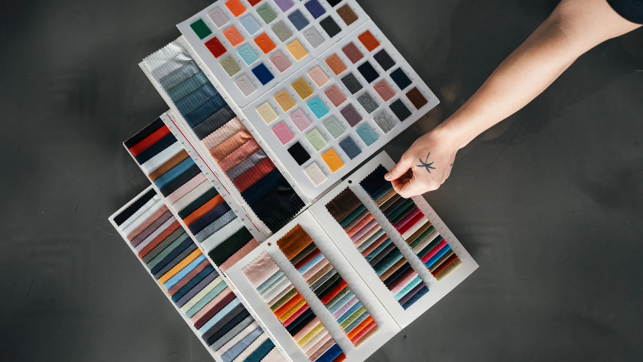 Person selecting fabric swatch from color palettes on a table, showcasing various shades and textures, with a hand tattoo visible.