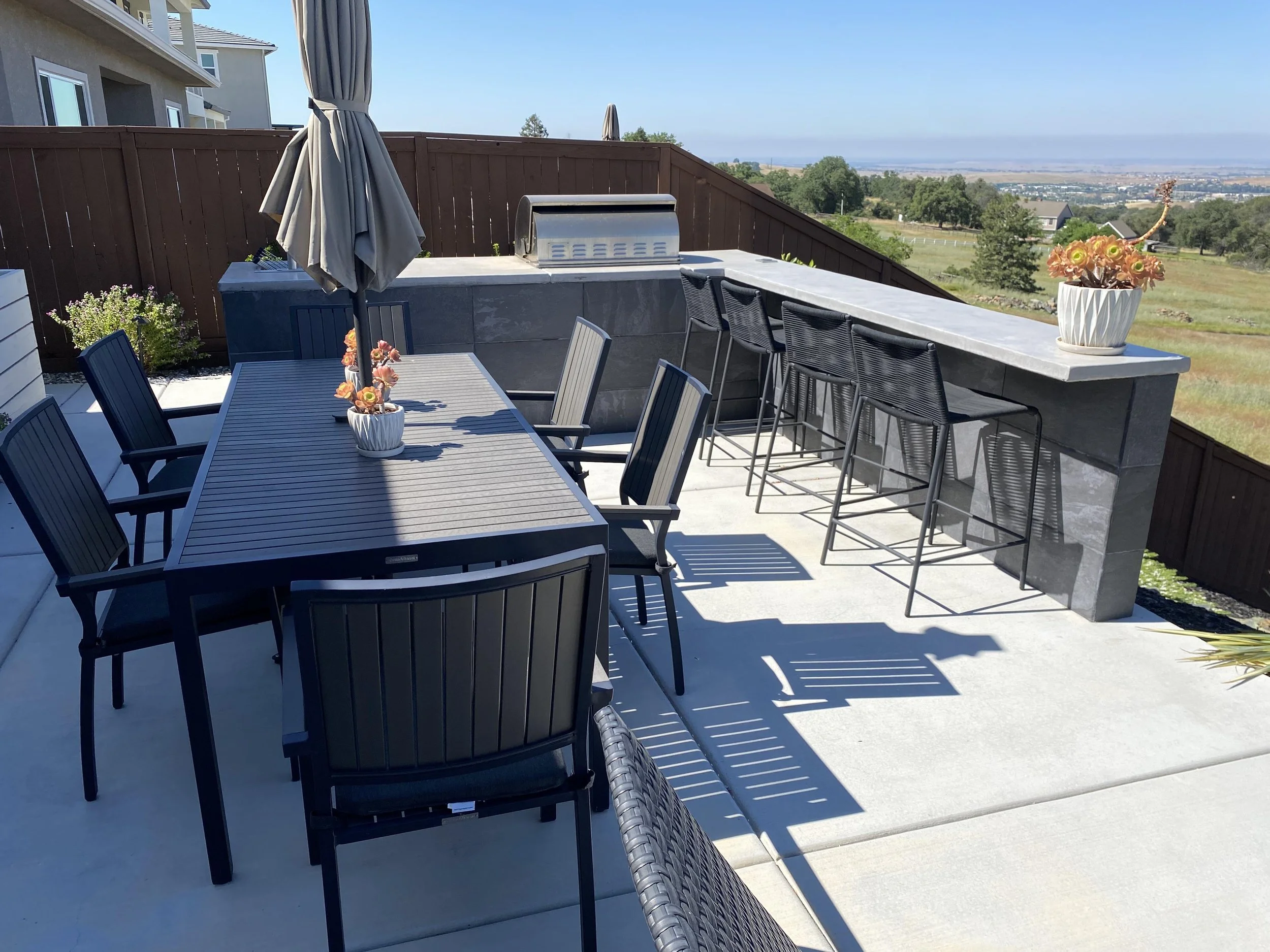 Outdoor patio with a dining table, chairs, bar stools, an umbrella, a grill, potted flowers, and scenic view.