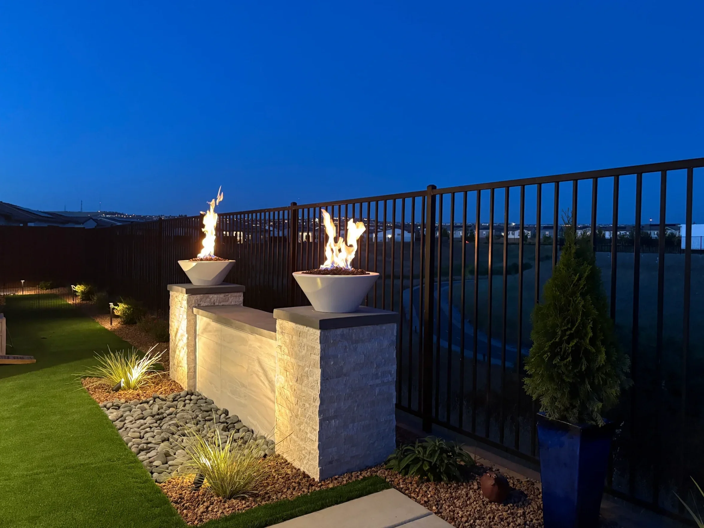 Outdoor backyard scene at dusk with fire bowls on a stone wall, illuminated green grass, small trees, and a metal fence beyond.