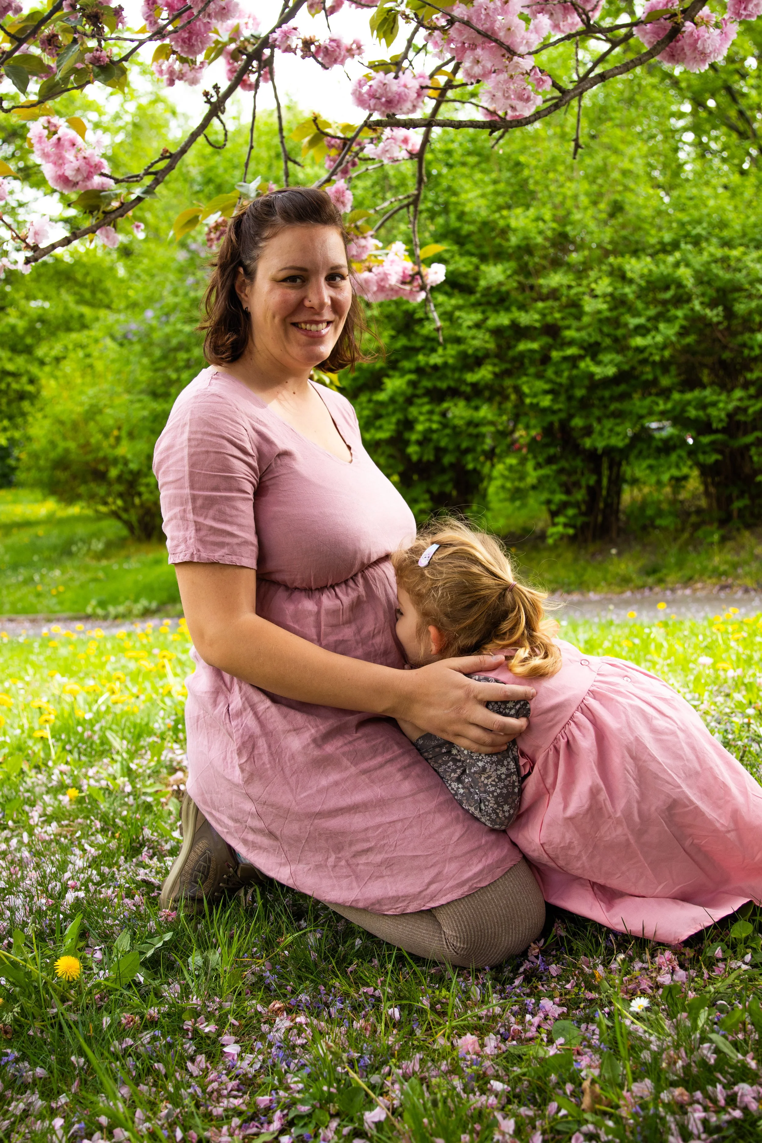 A woman and a young girl in pink dresses enjoying a moment in a park with blooming cherry blossoms and a carpet of colorful flowers.