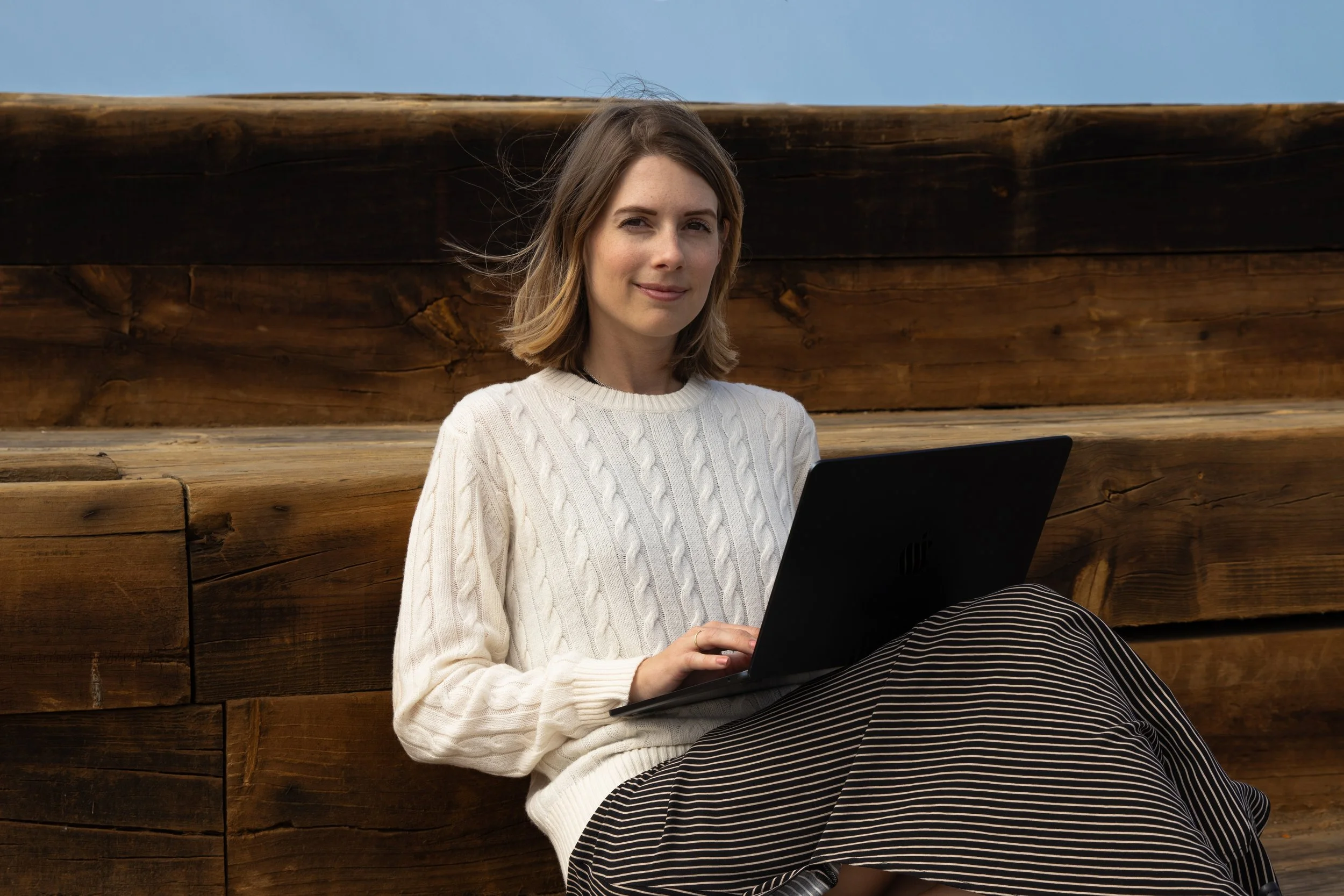 A woman with shoulder-length brown hair sitting outdoors on wooden steps, using a black laptop, wearing a white cable-knit sweater and a black and white striped skirt, under a blue sky.