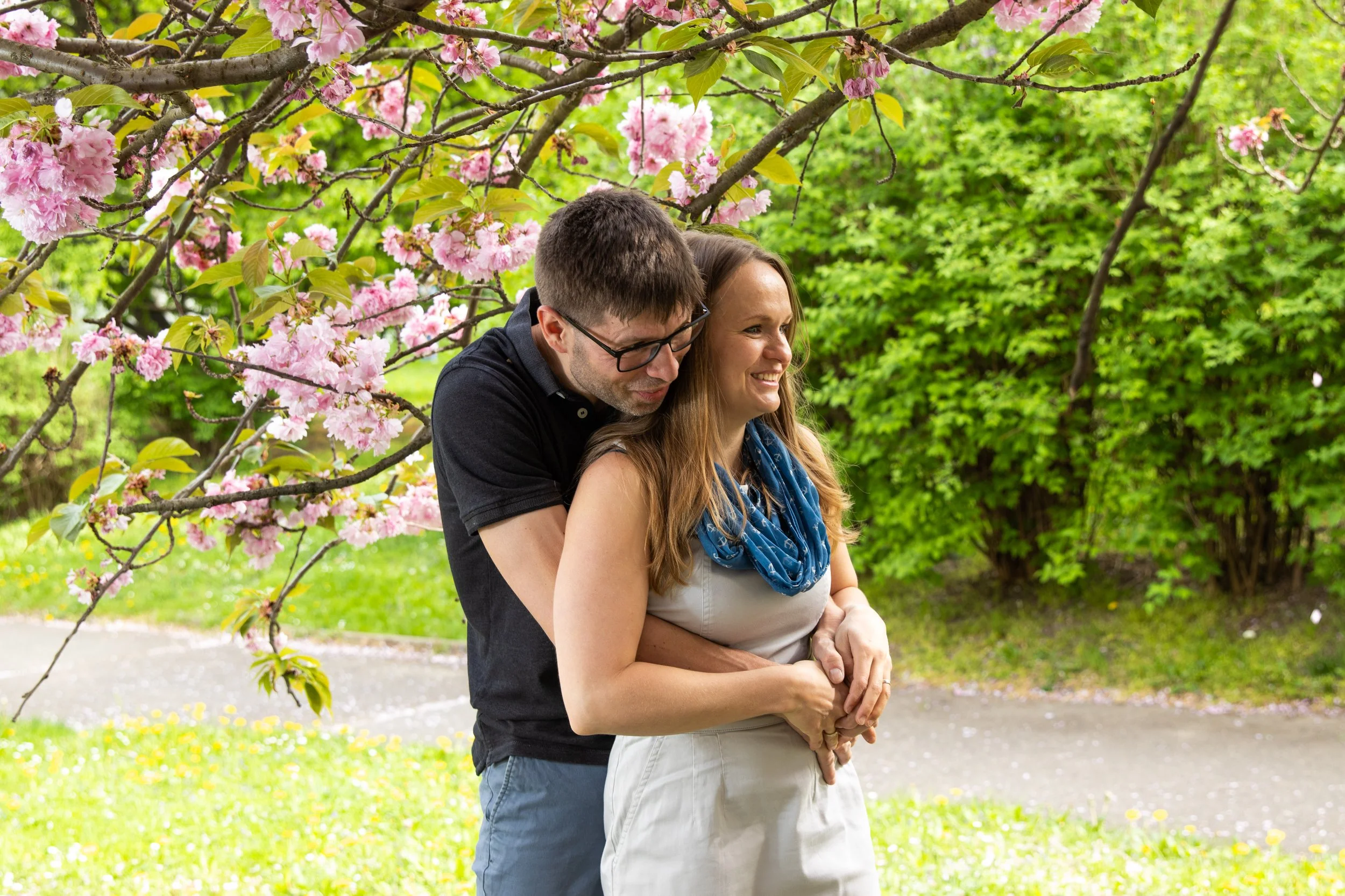 A man and woman embracing under a flowering pink tree in a park. The man has glasses and short dark hair, and the woman has long light brown hair and is smiling. There are green bushes and a sidewalk in the background.