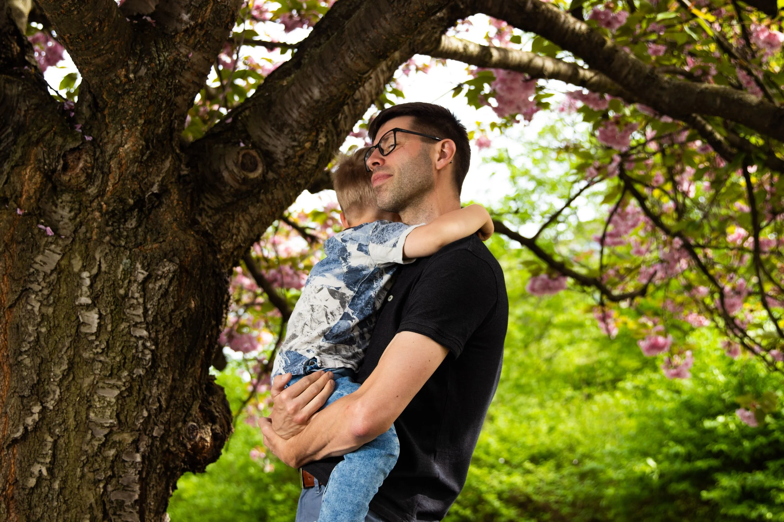 A man holding a young boy in embraces under a blooming pink cherry blossom tree, with green foliage in the background.