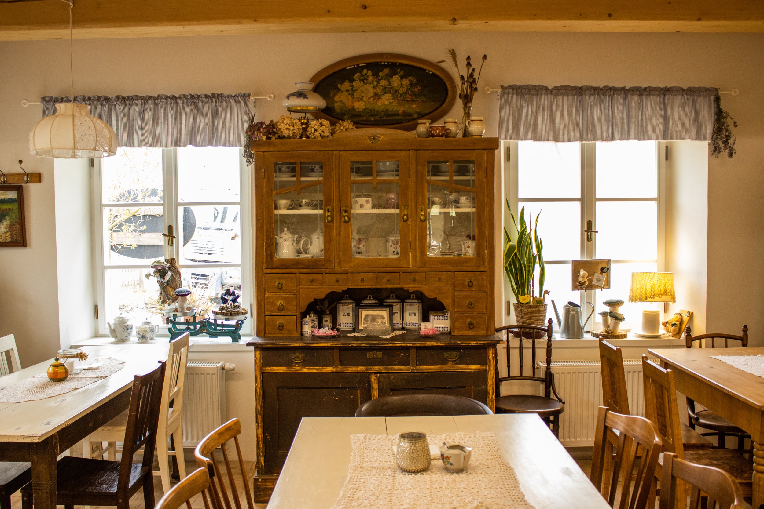 A cozy, vintage-style dining room with a large wooden hutch, multiple windows with curtains, and various decorative items including ceramics, plants, and small lamps on the tables and windowsills.