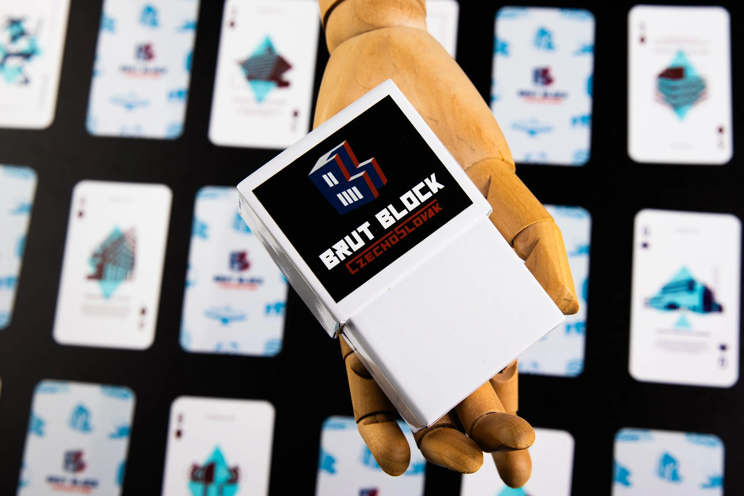 A wooden mannequin hand holding a box with the logo and text "Brut Block Czech Slovakia" in front of a background of playing cards laid out on a black surface.