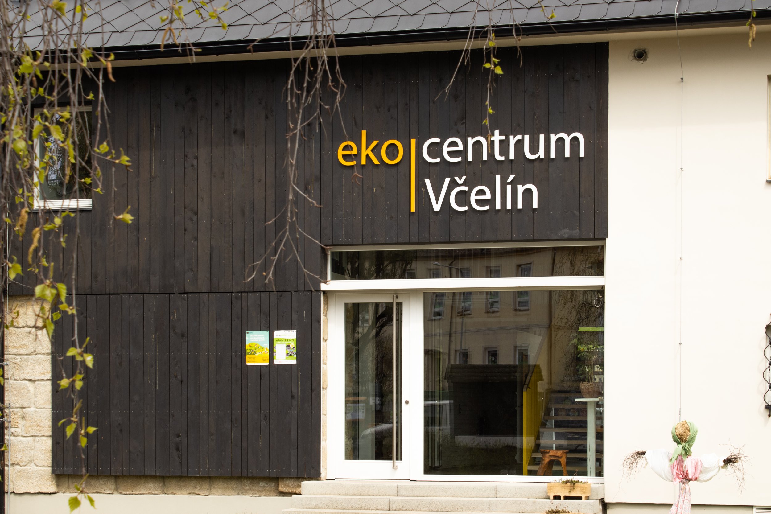 The exterior of a building with a sign that reads 'eko centrum Včelin' in white and yellow letters on a black wooden facade, with a glass door and some decorations outside.