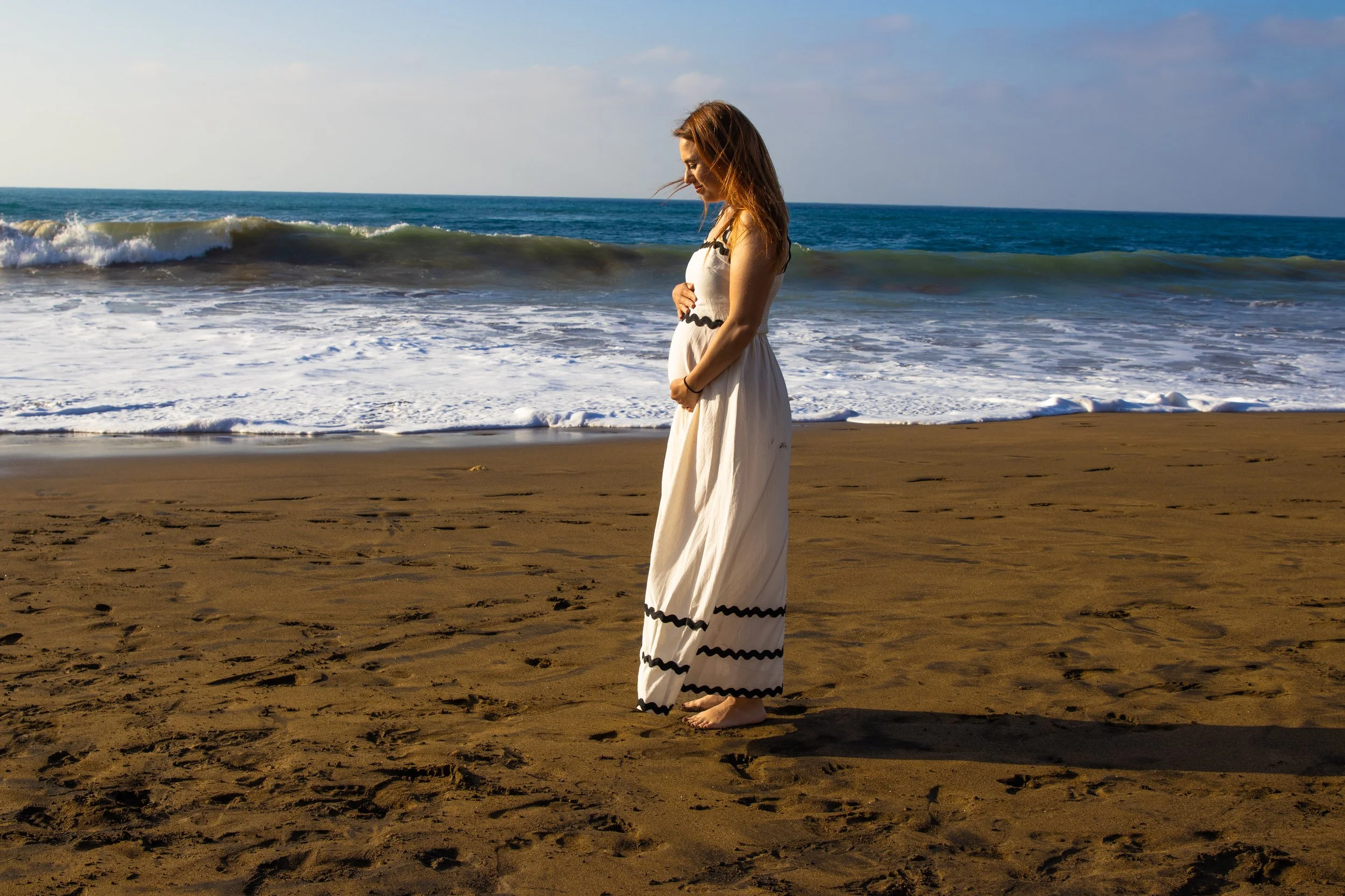 A woman standing barefoot on a sandy beach with the ocean and waves in the background, looking down thoughtfully.