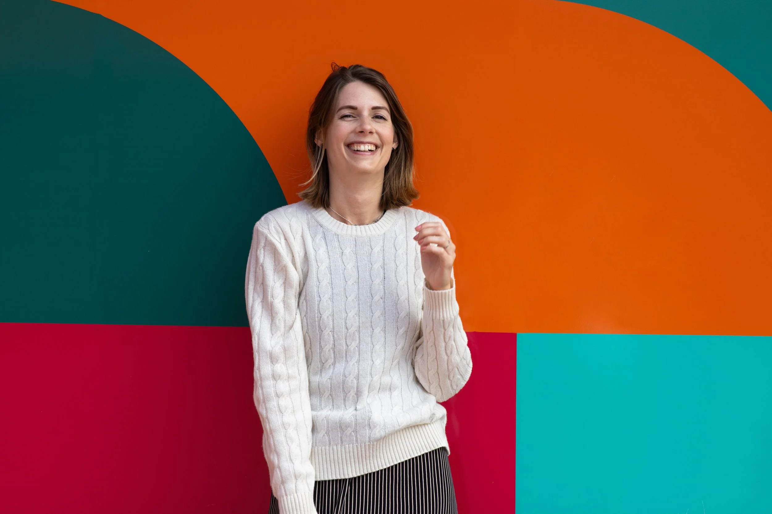 A woman with shoulder-length brown hair, wearing a white cable-knit sweater and striped pants, smiling and standing in front of a colorful, geometric background with orange, teal, and red sections.