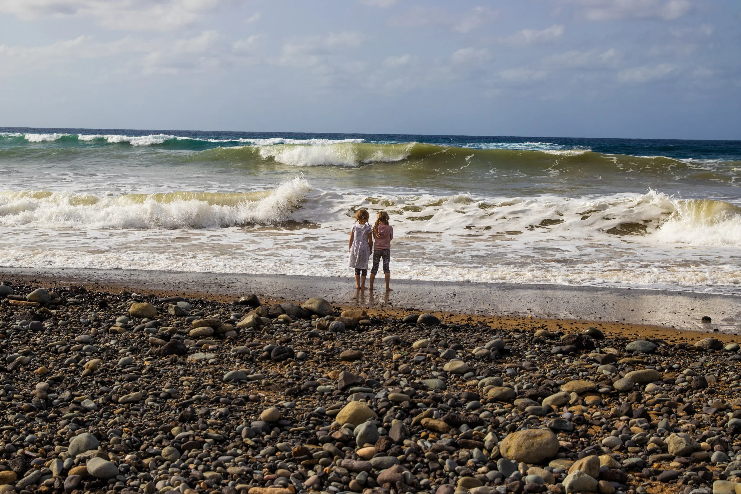 Two children standing ankle-deep in the ocean waves on a rocky beach, looking towards the horizon on a partly cloudy day.