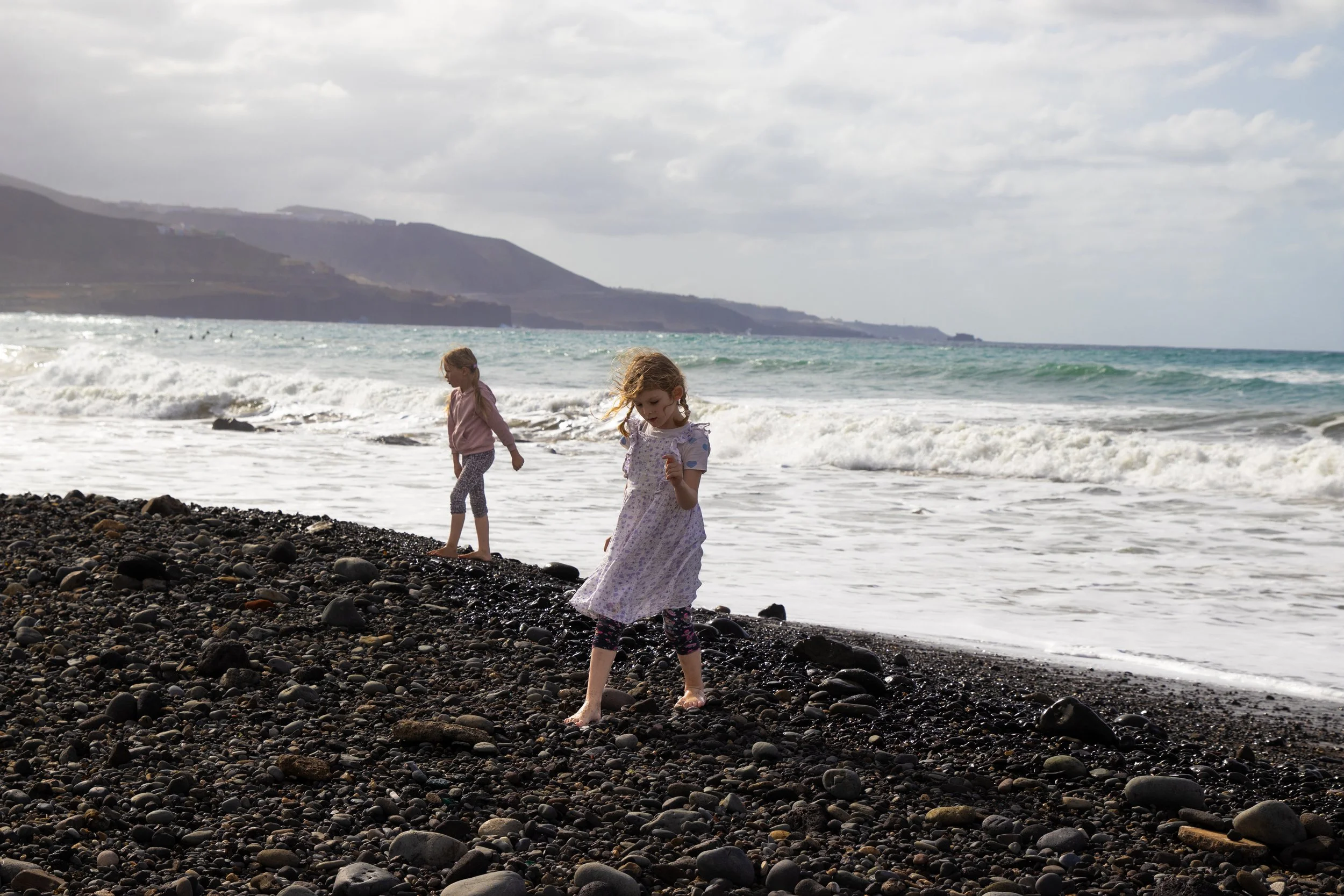 Two young girls walking barefoot on a rocky beach near the water's edge with waves and distant hills in the background.