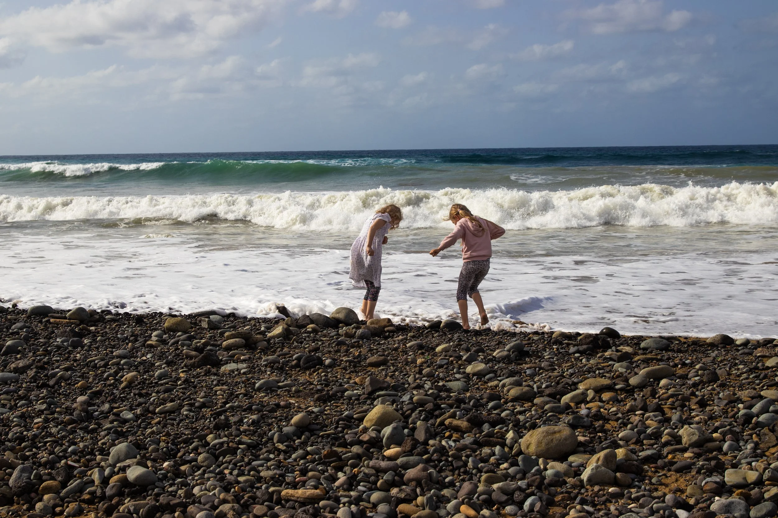 Two young girls playing near the shoreline on a rocky beach with ocean waves and clouds in the sky.