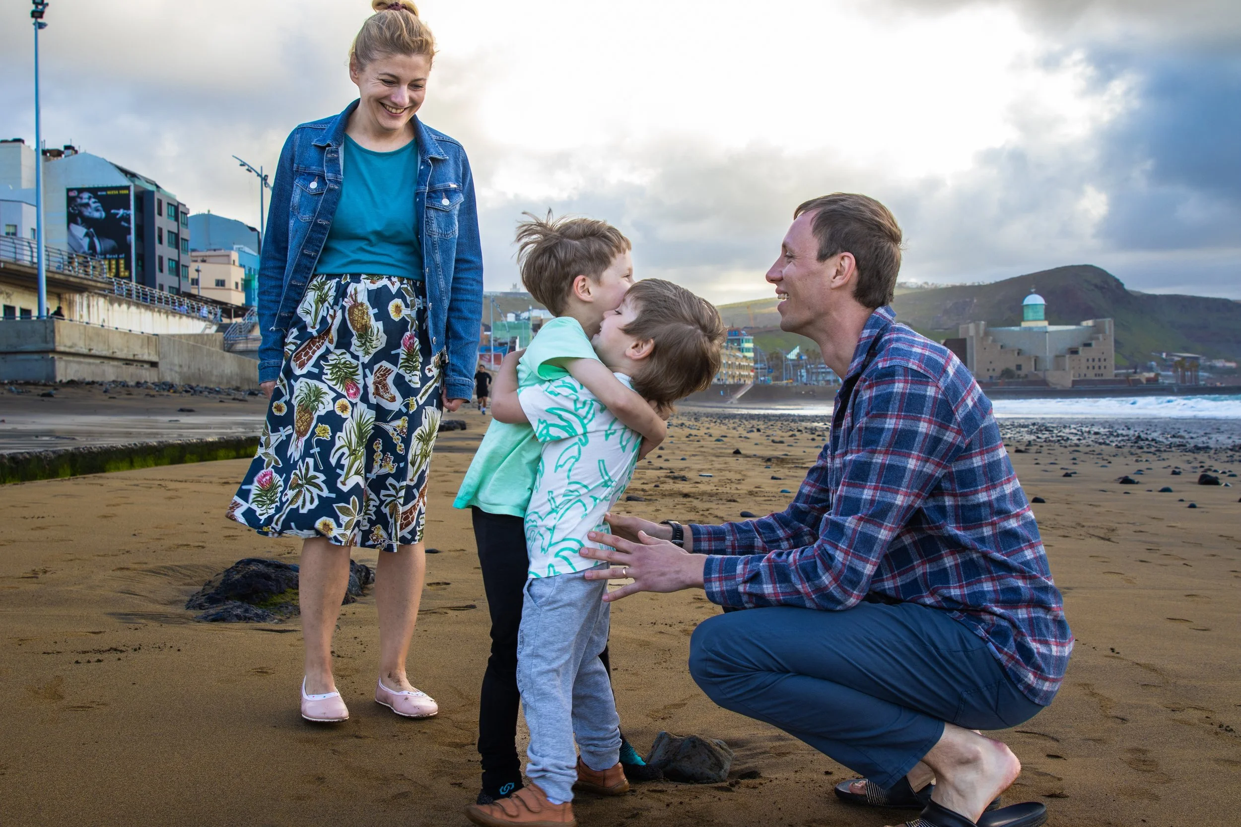 A family enjoying time on the beach with two children and two adults, one man kneeling and holding a young boy, and one woman standing, all smiling.