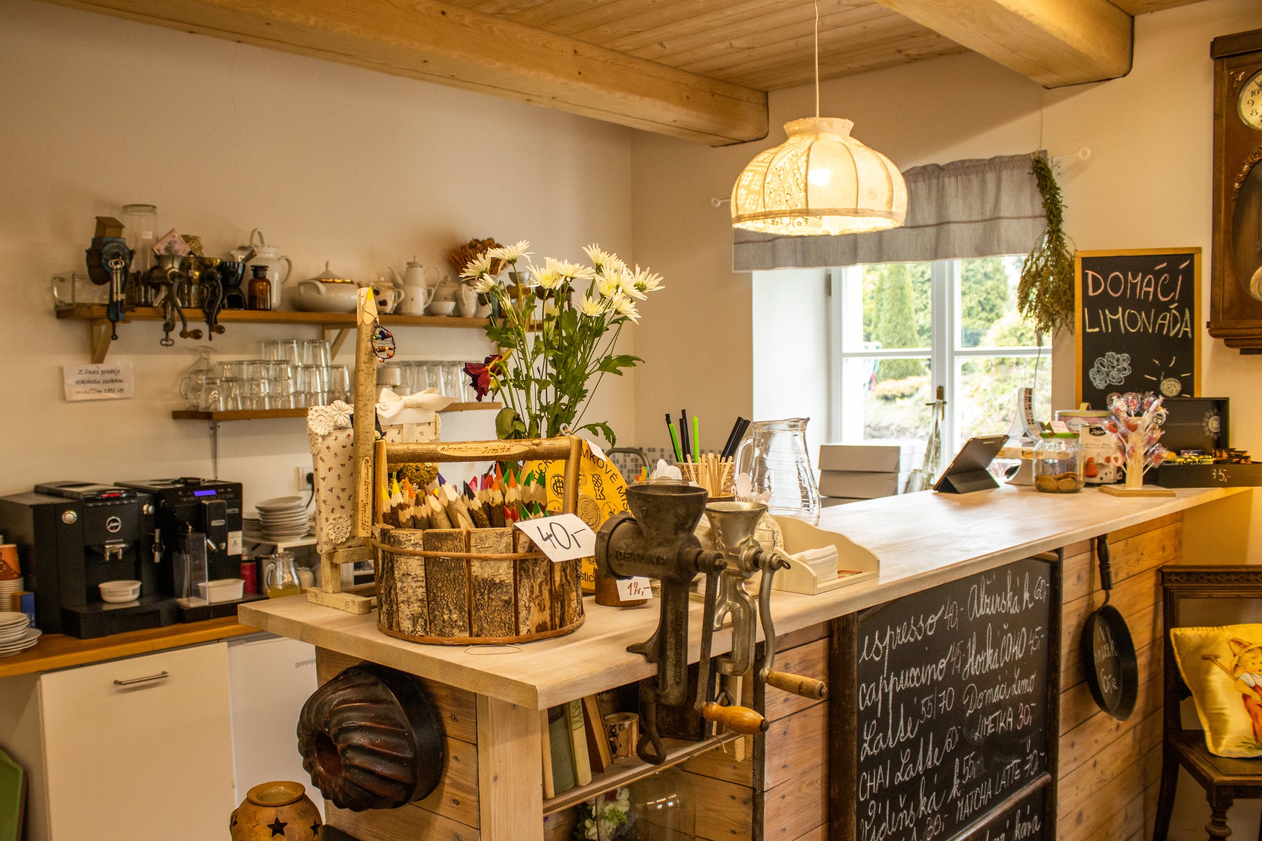 Interior of a cozy cafe with a wooden counter, flowers, a chalkboard menu, coffee equipment, and a window with natural light.