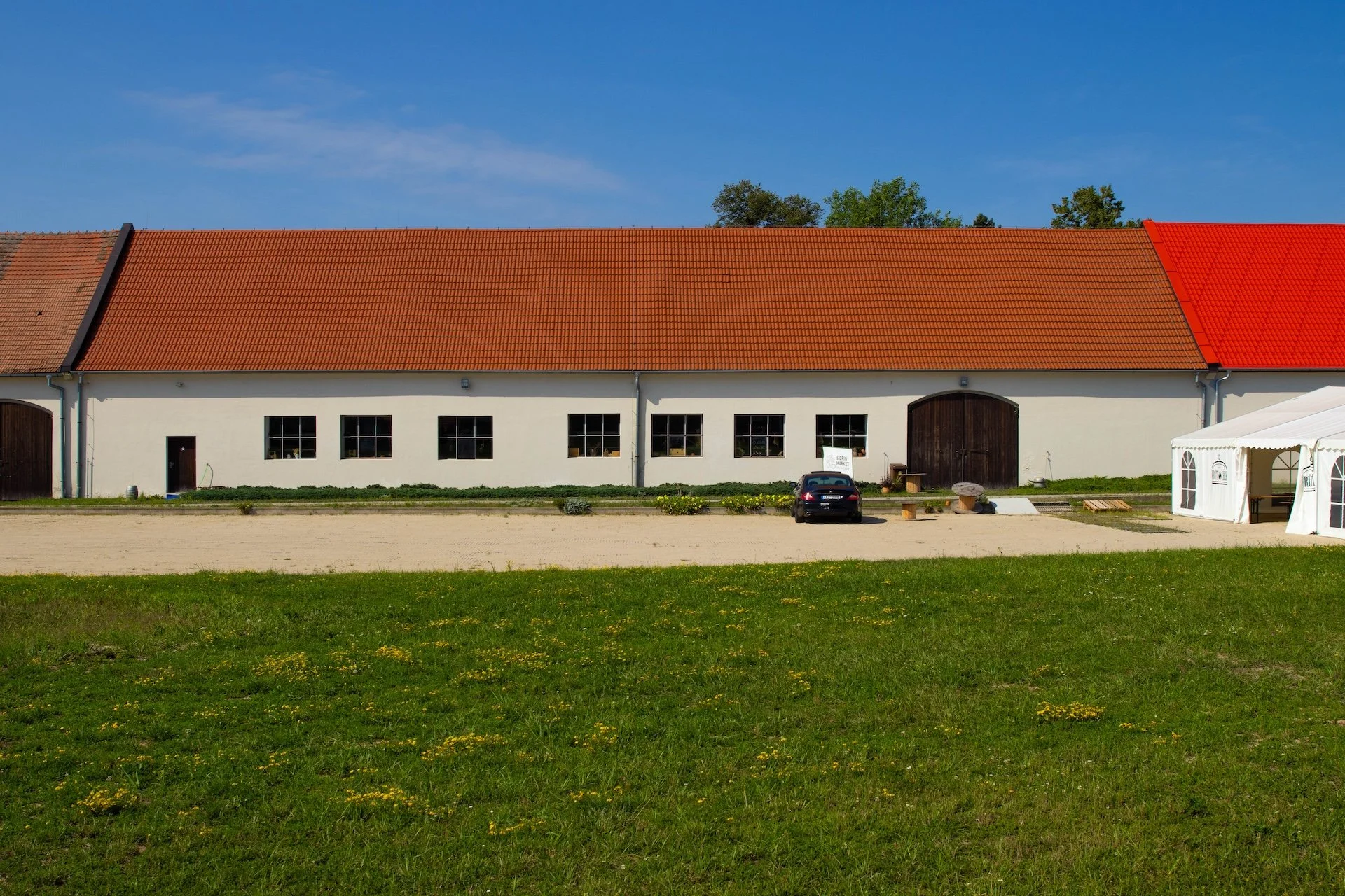 A large white barn with a red tiled roof on a farm, with a small black car parked in front, and a white tent on the right side, under a blue sky.