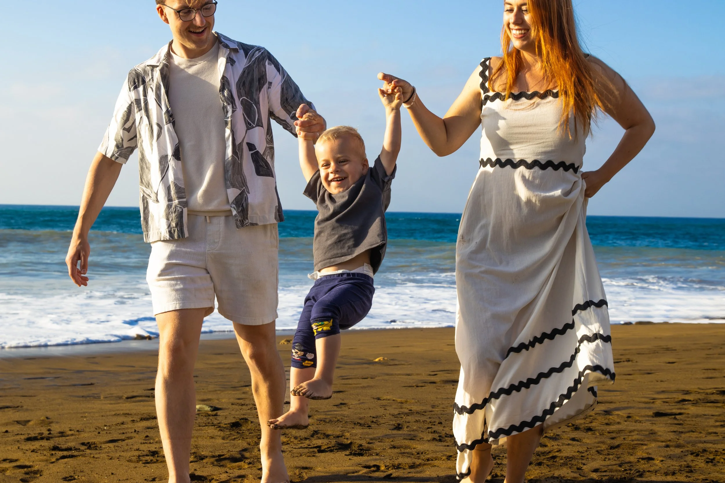 A happy family of three walking on the beach, with the father and mother holding hands and the young son swinging between them, smiling and enjoying the sunny day.