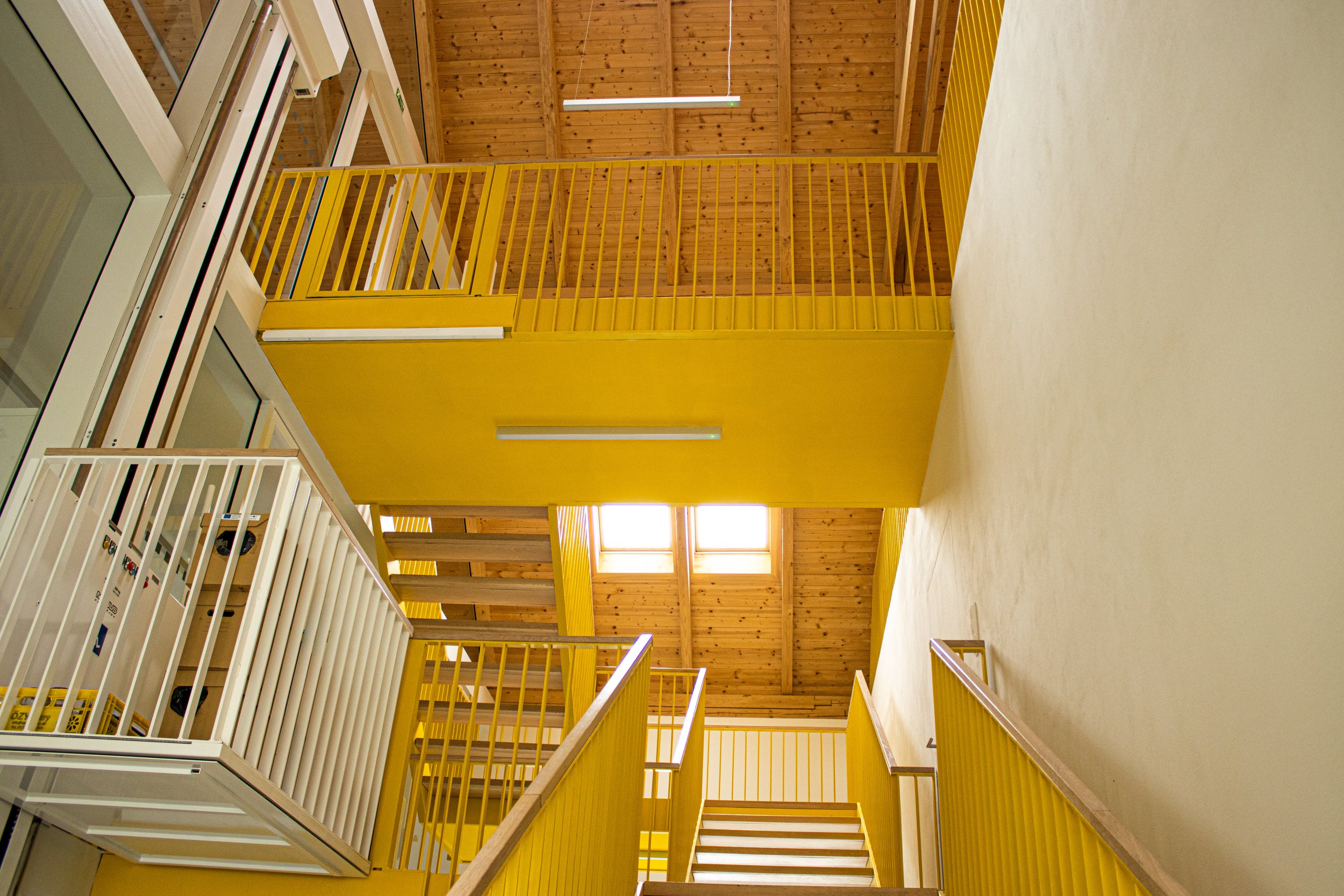 Interior view of a multi-story building with yellow railings, wooden ceilings, and staircases connecting different levels.