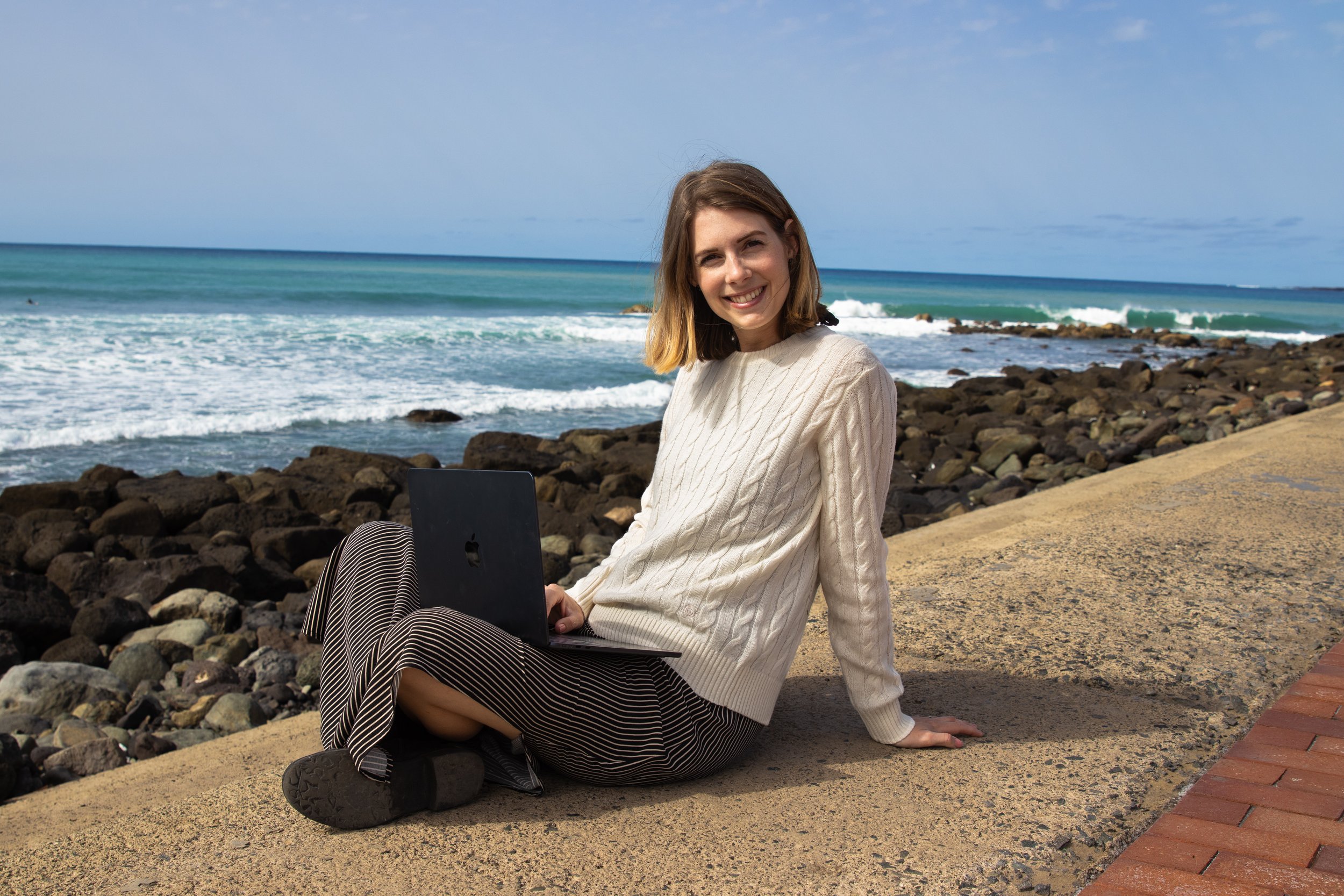 A young woman with shoulder-length blonde hair sitting on a concrete ledge by the rocky beach, holding a laptop, smiling at the camera with the ocean and sky in the background.