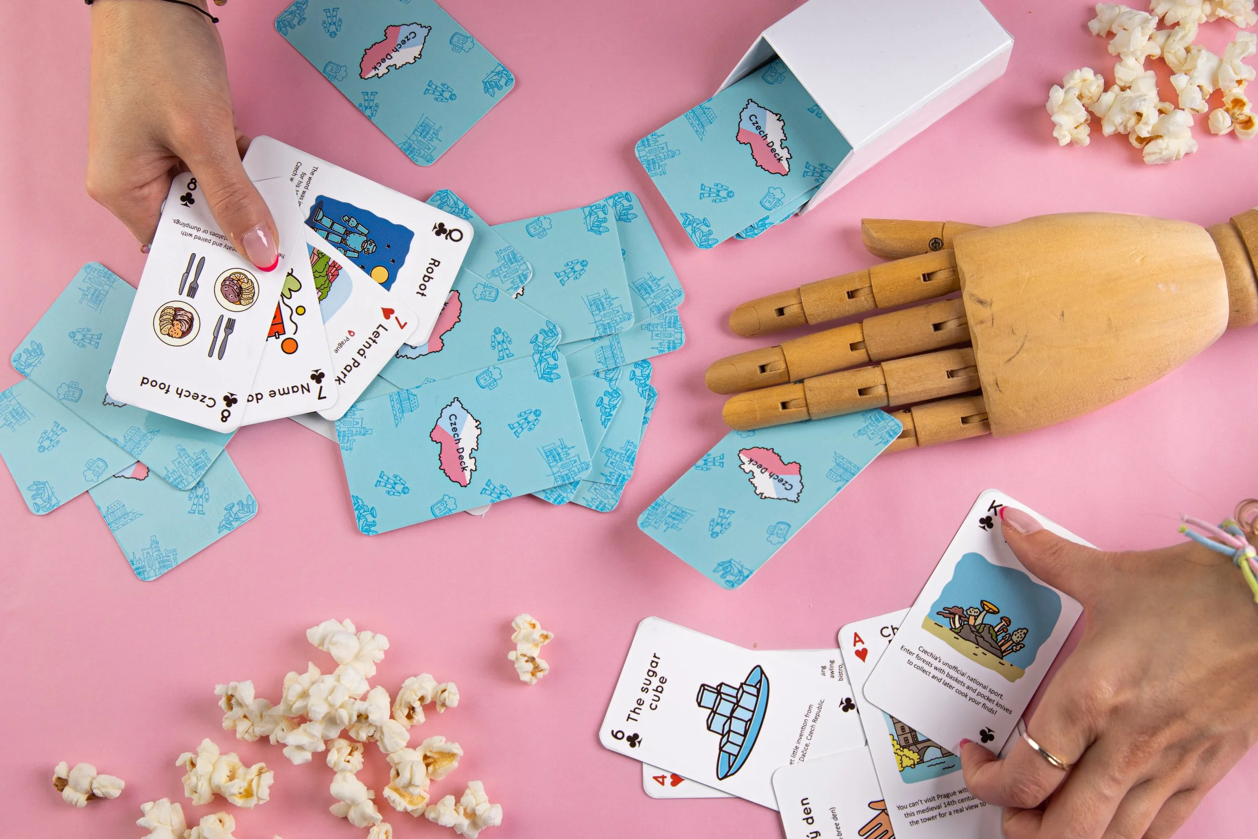 People playing a card game called Czechs in a pink space with popcorn, a wooden mannequin hand, and a cardboard box. The cards are themed with illustrations and text about Czech landmarks and food.