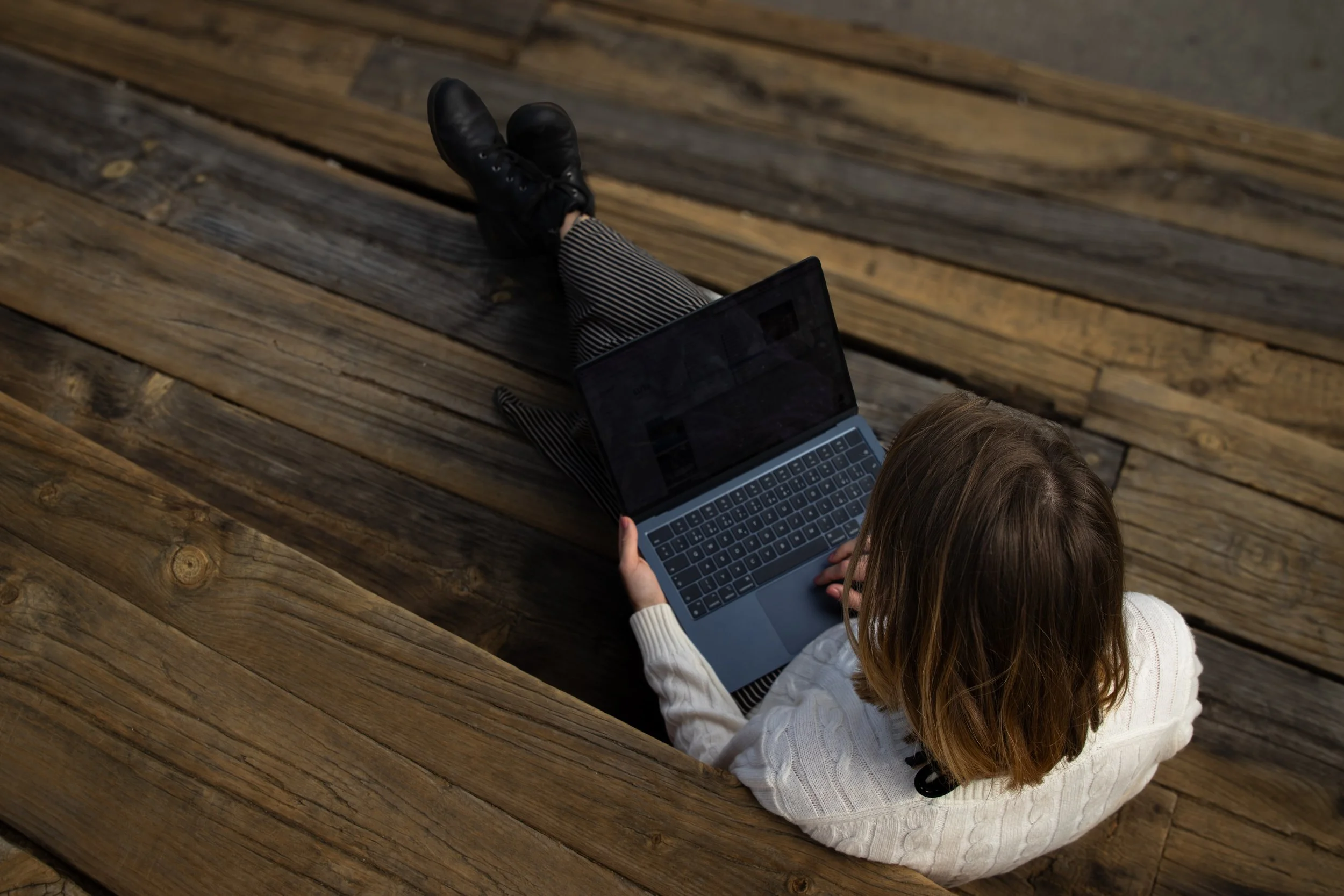 Young woman sitting outdoors on wooden benches, using a laptop. She has shoulder-length brown hair and is wearing a white sweater, striped pants, and black shoes.