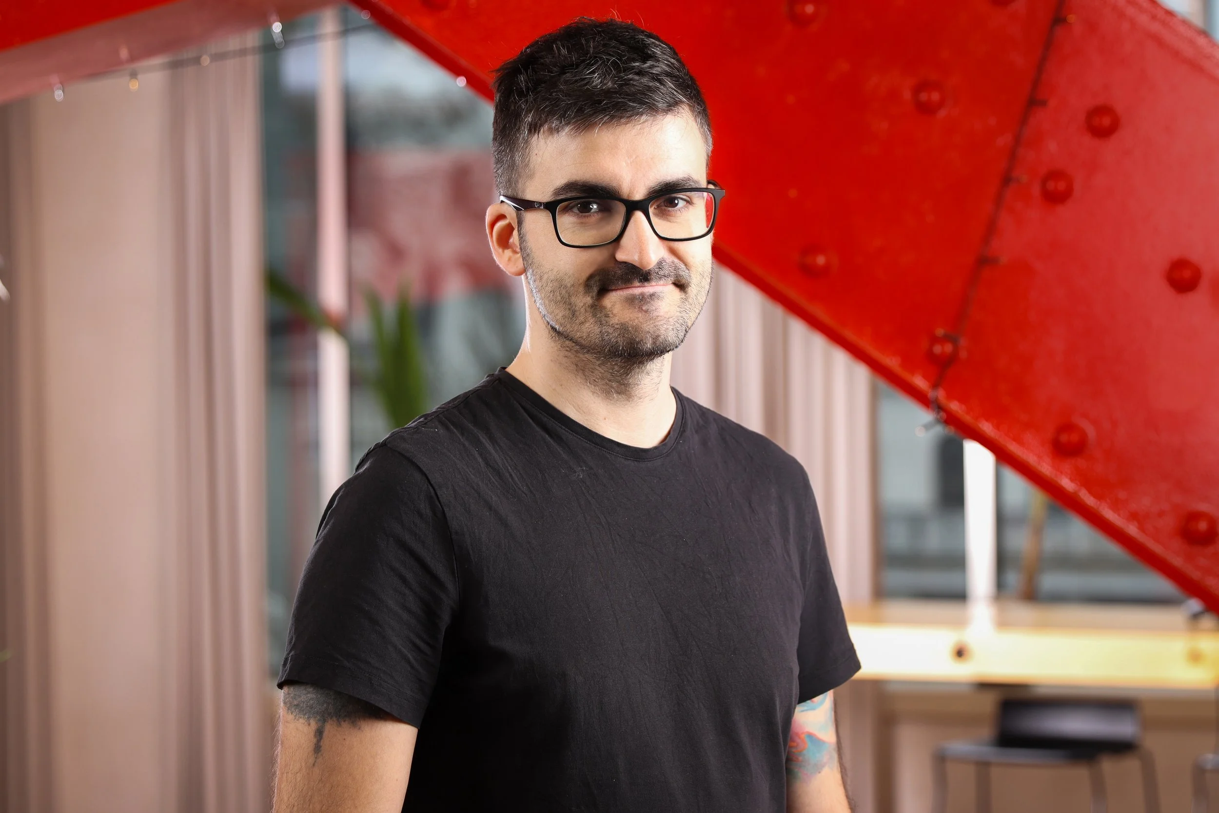 A man with short dark hair, glasses, and a beard wearing a black t-shirt standing indoors with a red structure in the background.