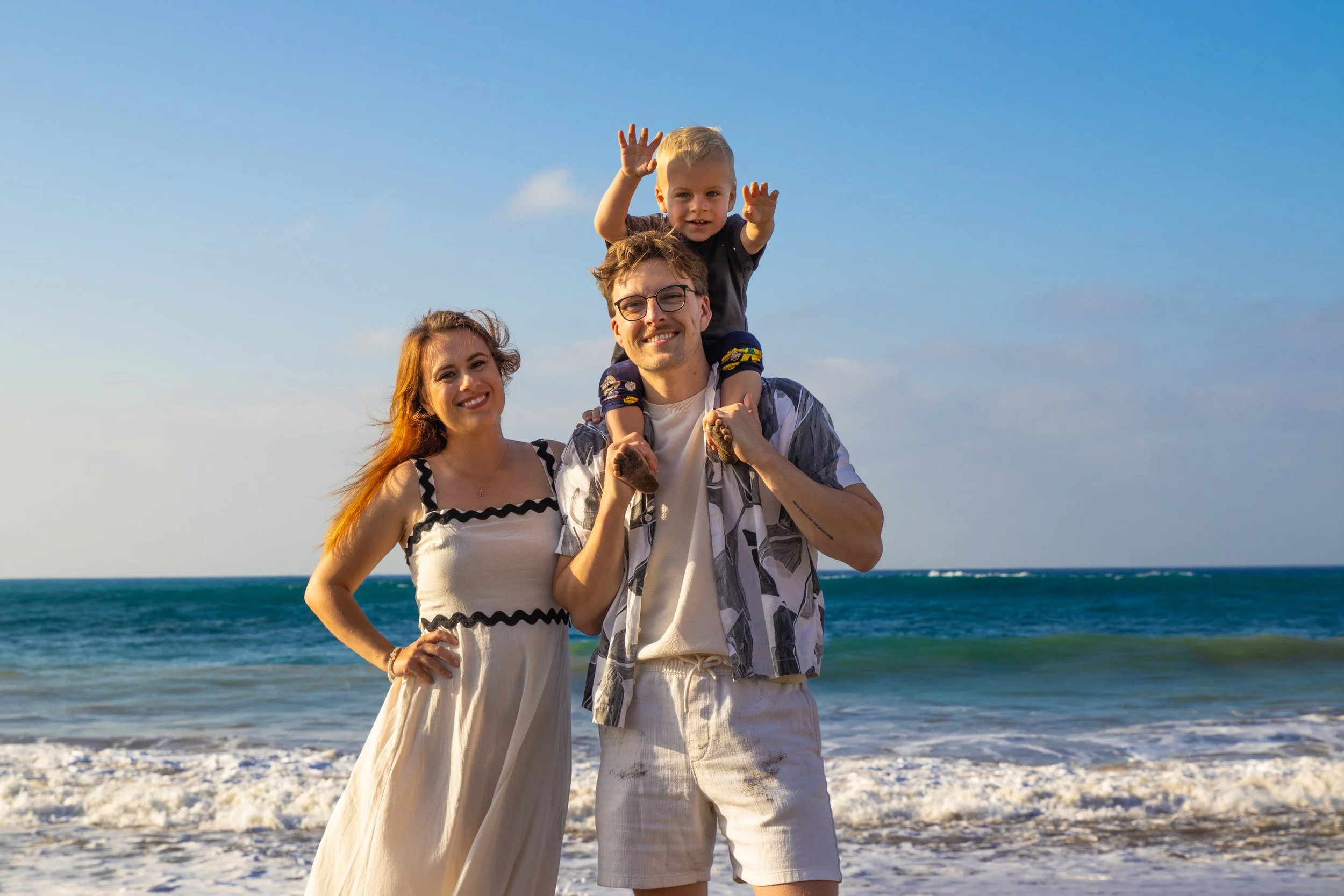 A happy family of three enjoying the beach with blue skies, a woman in a white dress, a man in a patterned shirt and beige shorts, and a young child on the man's shoulders waving.